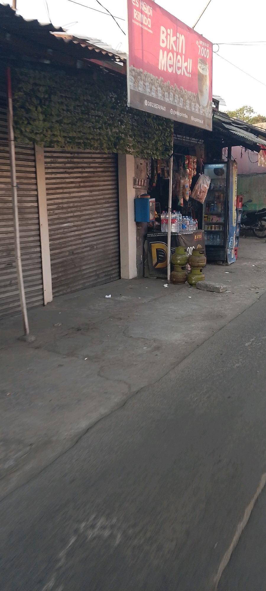 The image shows a small storefront in Karangsatria, Indonesia. The store appears to be a convenience store or a small food stall, selling drinks and possibly snacks. A prominent red sign hangs above the storefront, advertising "ES KOPI RIMBA - BIKIN MELEK!!" which translates to "JUNGLE COFFEE - MAKES YOU AWAKE!!". The sign also indicates a price of Rp 7.000 and displays an image of a coffee drink.
The storefront itself has a corrugated metal roll-up door that is partially open, revealing shelves stocked with bottled drinks and bags of snacks. To the right of the entrance, there is a blue dispenser, possibly for water or a vending machine. In front of the store, stacked on the ground, are several green propane gas cylinders. A blue and white refrigerator is visible behind the counter, filled with beverages.
The ground in front of the store is paved with concrete, and there are some scattered pieces of trash. The building has a corrugated metal roof and is partially covered with green leafy vines on the left side. A motorcycle is parked to the right of the store, partially obscured by the building. The overall environment suggests a street-side establishment in a residential or commercial area. The lighting indicates it is daytime, possibly late afternoon or early evening.