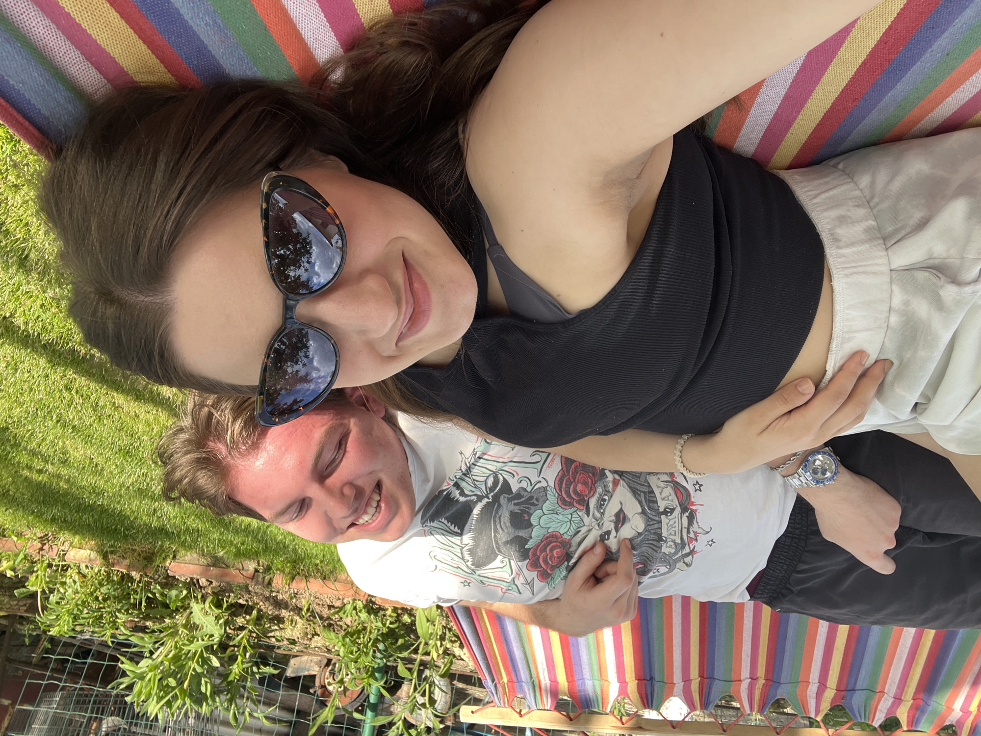 Two individuals are lying in a striped hammock in an outdoor setting. The woman is positioned above the man and is wearing sunglasses, a black top, and white shorts. The man is wearing a white t-shirt with a graphic print and black shorts. The surrounding environment includes green grass, plants, and a brick structure. The location appears to be in a garden setting, likely within the vicinity of Busmantsi, Bulgaria.
