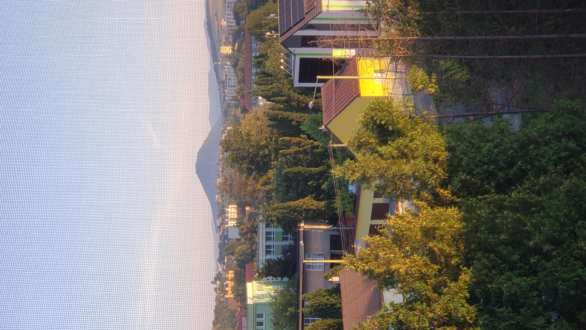 A telephoto shot captures a city view in Prešov, Slovakia, with a mesh screen in the foreground distorting the image slightly. The scene is bathed in warm, late afternoon light. In the middle ground, various buildings are visible, including a yellow residential building with brown garage doors, a beige building with garages and windows, and a longer, light-colored building with a pitched roof and dark windows. Behind these, a cluster of trees, some with golden foliage, dominate the landscape. Further in the distance, more buildings, including apartment blocks and older structures with light-colored facades and red roofs, are scattered amongst green trees. A prominent conical hill with some greenery on its slopes rises in the background, silhouetted against a hazy, light blue sky. The overall impression is one of a suburban or semi-urban environment on a clear day.