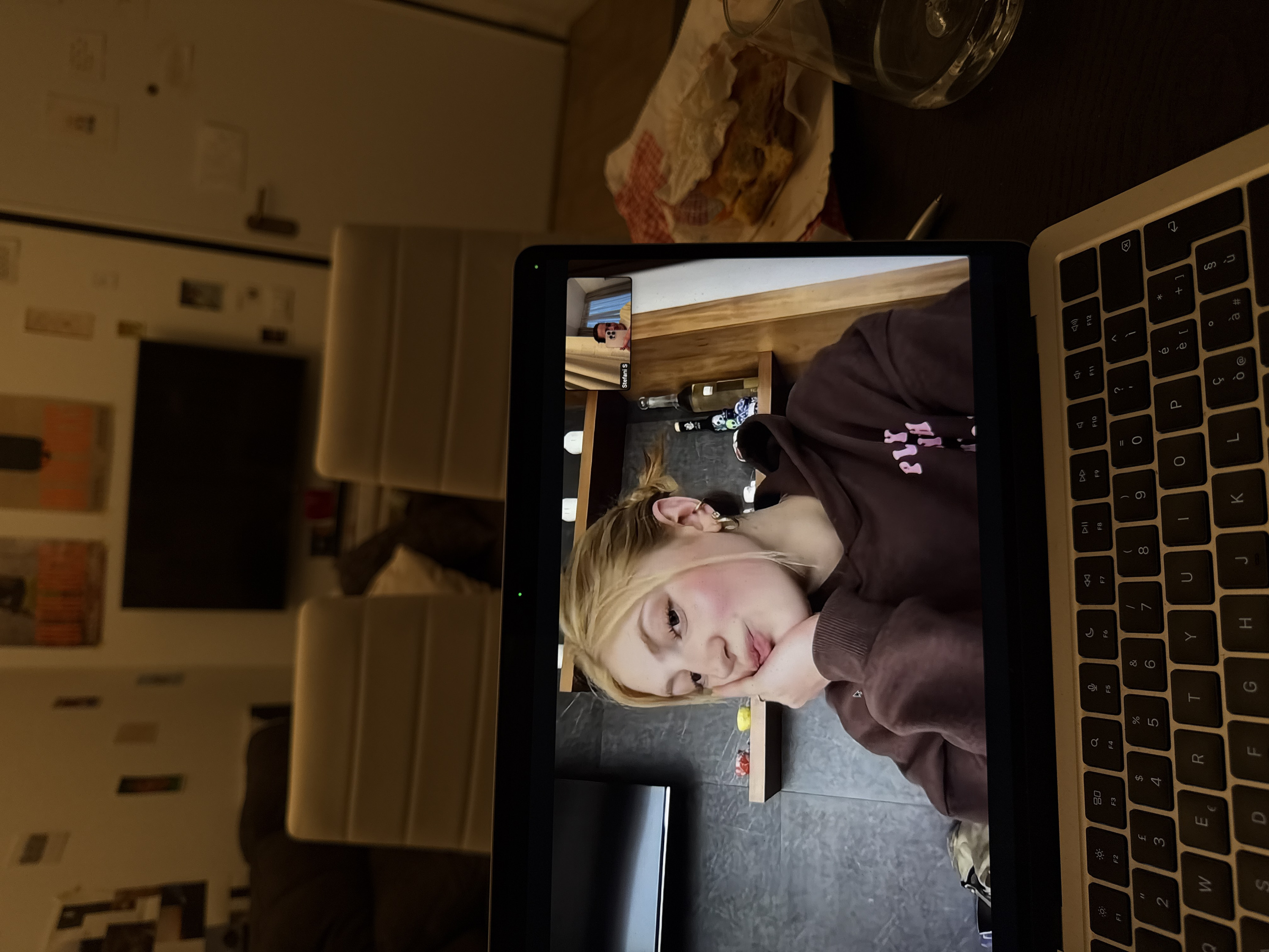 The image displays an indoor scene, likely a residential interior in Milan, Italy, captured from a first-person perspective. The foreground features an open silver-colored laptop, prominently showing its keyboard with a QWERTY layout including Italian-specific characters (e.g., '€' on the 'E' key, 'ù' on the 'P' key, 'ç' on the 'L' key). To the right of the laptop, on a dark tabletop, rests a clear glass containing water, a silver pen, and a partially consumed pastry or dessert item wrapped in white paper with red printed details.
The laptop screen displays an active video call. The main window features a young blonde female, with her chin resting on her right hand, looking slightly away from the camera with a subtle expression. She is wearing a dark brown hooded sweatshirt with light pink text. Her background on the video call shows a dark, possibly textured wall with two wooden shelves holding various items, including small colorful objects and two bottles. In the top-right corner of the video call interface, a smaller thumbnail shows a second participant, identified by the name "Stefani S," holding a mobile phone. A small green indicator light is visible on the laptop's bezel above the main video feed.
The physical background of the room is dimly lit with warm light. A large, dark flat-screen television is mounted on a light-colored wall. The walls surrounding the television and a door on the far right are sparsely decorated with various papers or posters. Two light-colored, high-backed chairs are visible behind the laptop, positioned in front of the television. A dark sofa or armchair is partially visible on the far left.
