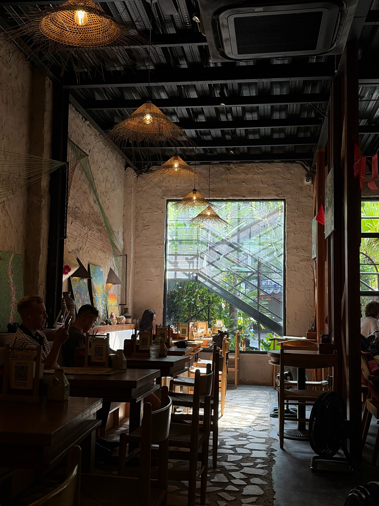 This is an interior shot of a restaurant in Da Nang, Vietnam. The restaurant has a rustic ambiance with exposed brick walls and wooden furniture. Natural light streams in from a large window, illuminating the dining area and highlighting the textured floor made of irregularly shaped stones. Several tables are set with placemats and small bottles, suggesting they are ready for or in the process of being served. People are seated at some of the tables, engaged in conversations or looking at their phones. Overhead, woven straw pendant lights cast a warm glow. In the background, a staircase is visible through the window, surrounded by lush greenery, indicating an outdoor or garden view. The ceiling is a dark industrial design with visible beams and ductwork, contrasting with the warmer elements of the restaurant's decor. A black standing fan is positioned near a table in the foreground.