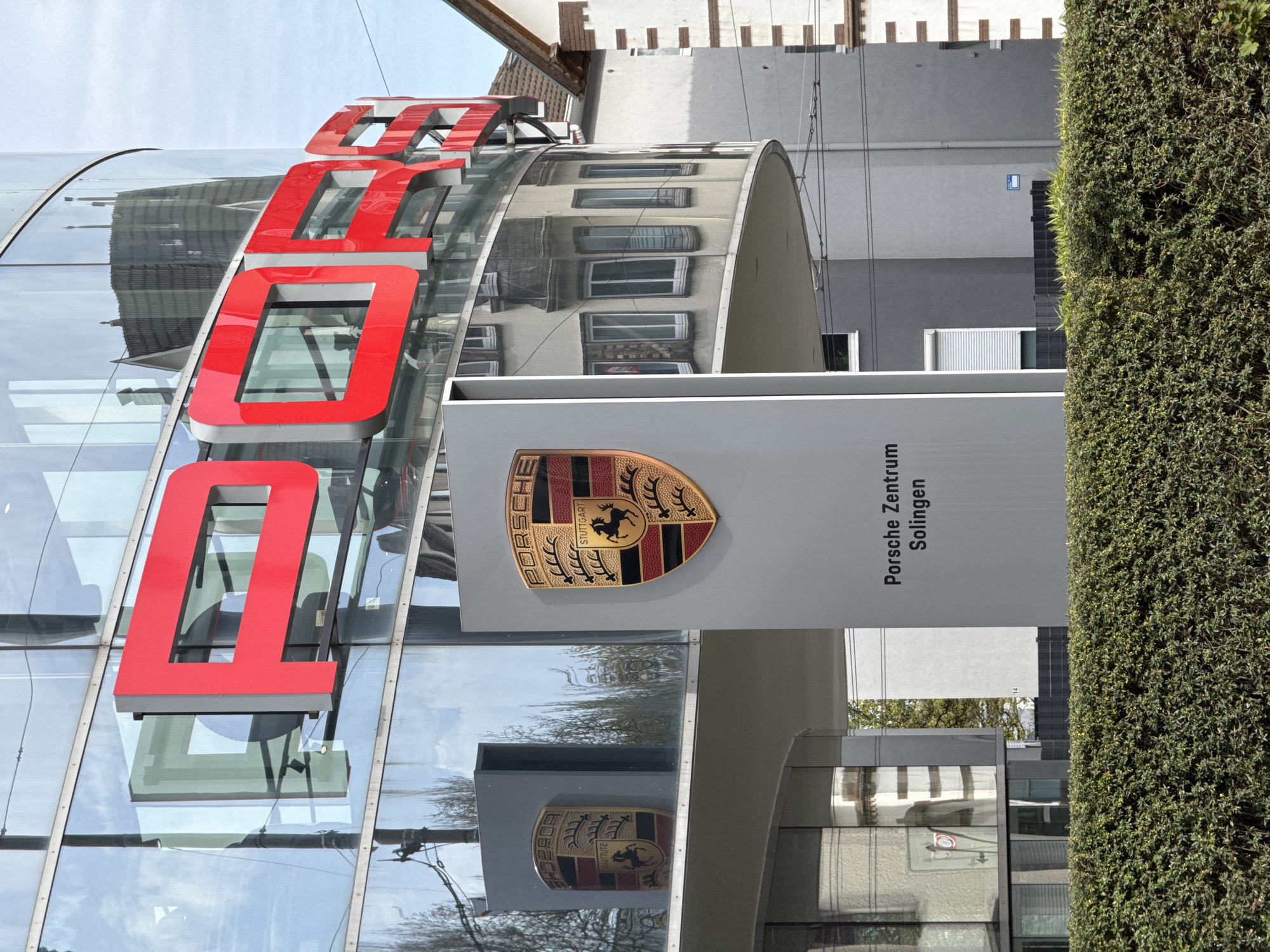 The image depicts the Porsche Zentrum Solingen, a car dealership located in Solingen, Germany. Prominent features include the dealership's modern glass facade with large, red "PORSCHE" lettering. A large, white sign with the Porsche emblem and "Porsche Zentrum Solingen" written vertically stands in front of the building. The reflection of nearby buildings can be seen on the glass. A tall, dense hedge is visible on the right side of the frame. The sky is light blue with some clouds.