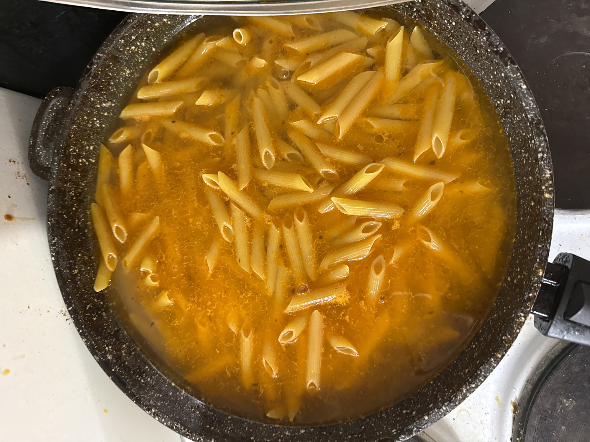 A top-down shot shows penne pasta in a broth-like liquid, cooking in a black speckled frying pan on a white stovetop. The stove is located in the city of Khimki, Russia.