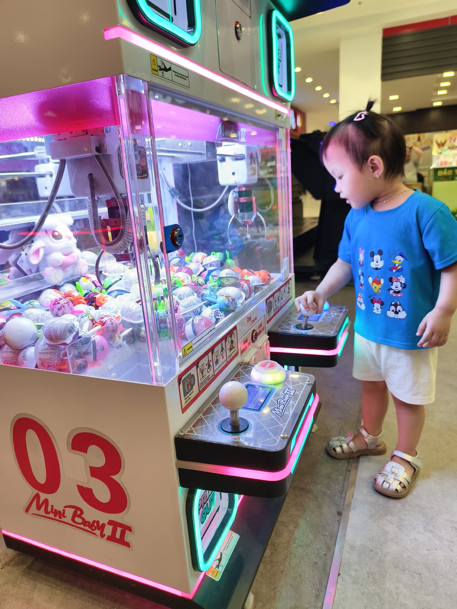 A young child is operating a "Mini Baby II" claw crane game, possibly in a shopping mall, located in the city of Unknown, Unknown. The game is brightly lit with pink and turquoise LED lights and filled with various toys and prizes, including plush animals and wrapped Kinder Surprise eggs. The child, wearing a blue T-shirt with cartoon characters and white shorts, stands near the game and touches the control panel. The flooring is tiled, suggesting an indoor commercial setting.