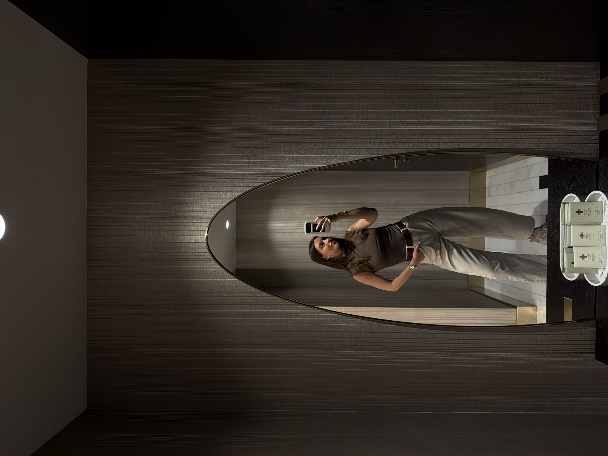 A vertically oriented medium shot depicts the reflection of a woman in an oval mirror. The woman holds a mobile phone as if taking a selfie. She is wearing a brown shirt, tan pants, and a belt. In the reflection, white-tiled walls can be seen, as well as small amenities on a black surface, contained in a white holder. The mirror is mounted on a wall with horizontal striped wallpaper. The lighting is ambient, creating soft shadows. The location could be inferred as a bathroom or dressing room in Budva, Montenegro, given the likely presence of amenities typically found in such spaces.
