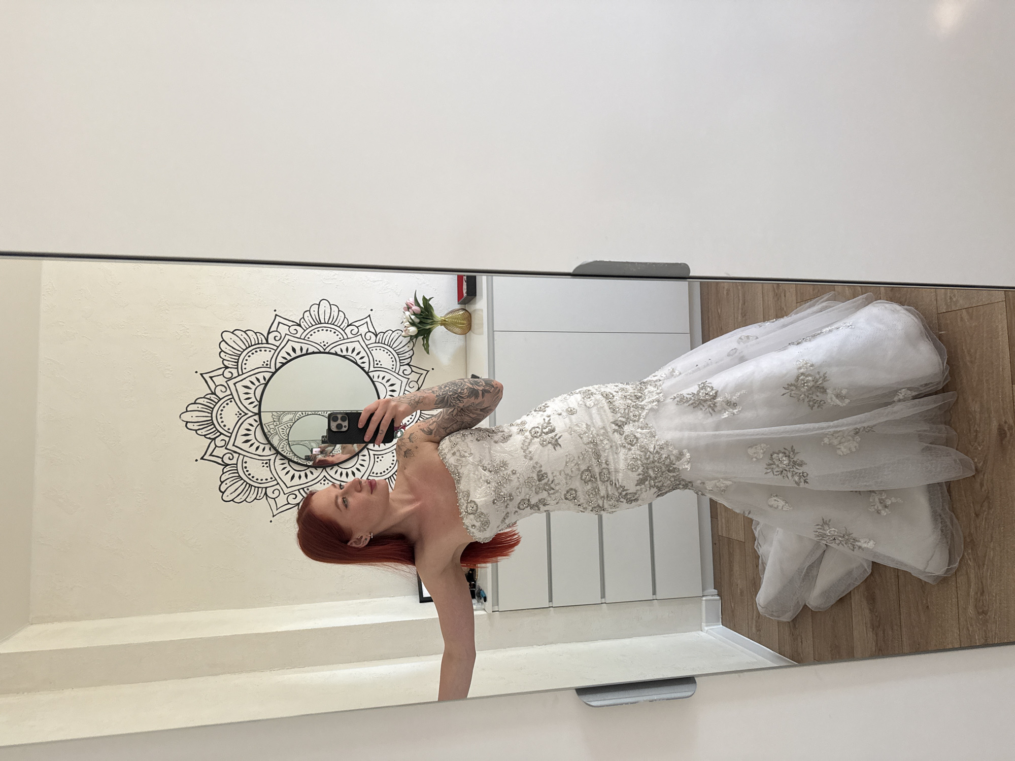 A female is captured in a full-length mirror selfie within an indoor residential setting, located in Rostov-on-Don, Russia. The subject, possessing long, straight red hair and visible tattoos on both arms, wears a strapless, white, mermaid-style gown. The dress is heavily embellished with silver and white floral embroidery and beadwork, particularly dense on the bodice and hips, gradually scattering down the sheer tulle skirt and train. She holds a black smartphone in her left hand, angled to capture her reflection, while her right arm rests against the frame of the mirror. Her head is slightly turned, and her gaze is directed towards the phone in the reflection.
The background, partially visible through the mirror, features an off-white, textured wall. A large, black, intricate mandala-style mural is painted onto this wall, with a circular mirror mounted centrally within its design. Below the mandala and mirror, a white dresser with multiple drawers is visible. On its surface, to the right, are a small vase containing pink tulips, a gold-colored vase, a small red box, and a black square box. To the viewer's left of the full-length mirror, a white wall section exhibits vertical decorative ridges. The flooring consists of light-colored wooden laminate planks. To the right of the full-length mirror, a white wardrobe door with a dark grey handle is present.