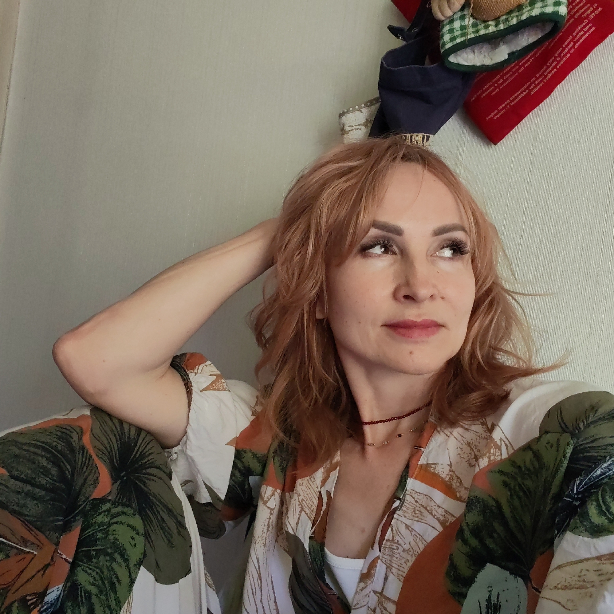 A fair-skinned woman with shoulder-length reddish-blonde hair is resting with her hand behind her head and looking upwards to the right of the frame. She is wearing a light-colored floral-patterned blouse. A red beaded necklace is visible around her neck. Above her head, there are some objects hanging on a wall, including what appears to be a small straw-filled figure and a red stocking-like object with white text on it. The background is a light-colored wall.
