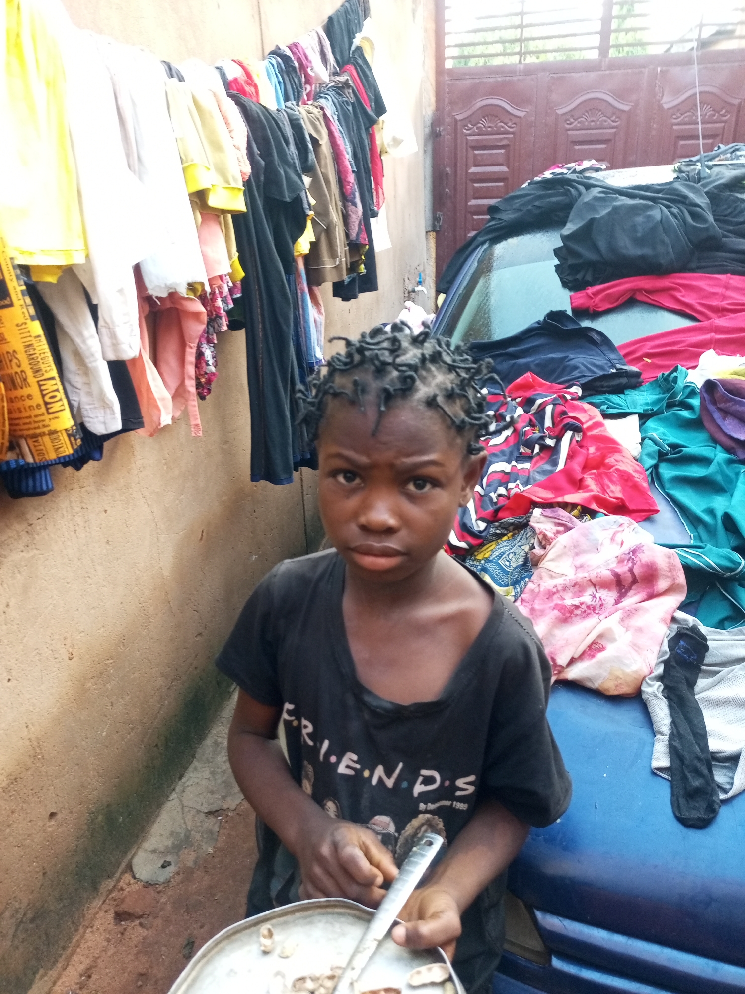 In Gombe, Nigeria, a young person with braided hair stands in front of a wall where laundry is hung to dry. The individual wears a black t-shirt with "F.R.I.E.N.D.S." printed on it and holds a metal plate with food. Behind them, clothes are draped over a blue car, and a metal gate is visible in the background. The scene appears to be an outdoor domestic setting.