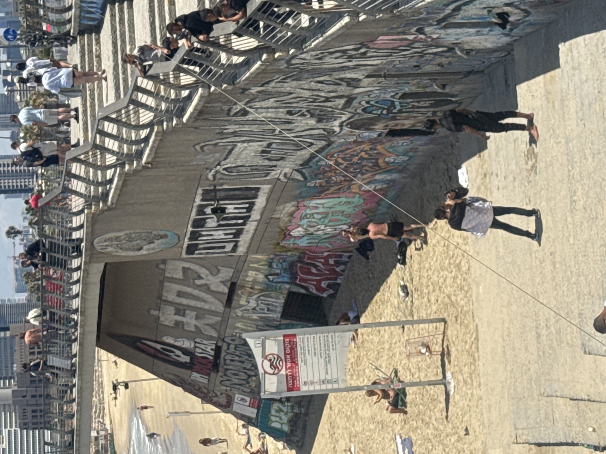 The image shows a sunny day at a beach in Tel Aviv, Israel, near a concrete overpass with graffiti. The graffiti includes writing in Hebrew and English. People are walking and standing on the bridge and beach. Some people are sunbathing on the sand. There's a sign with a red circle with a line through it. A cable runs diagonally through the scene. In the background are buildings in the city. The sea is visible in the distance.