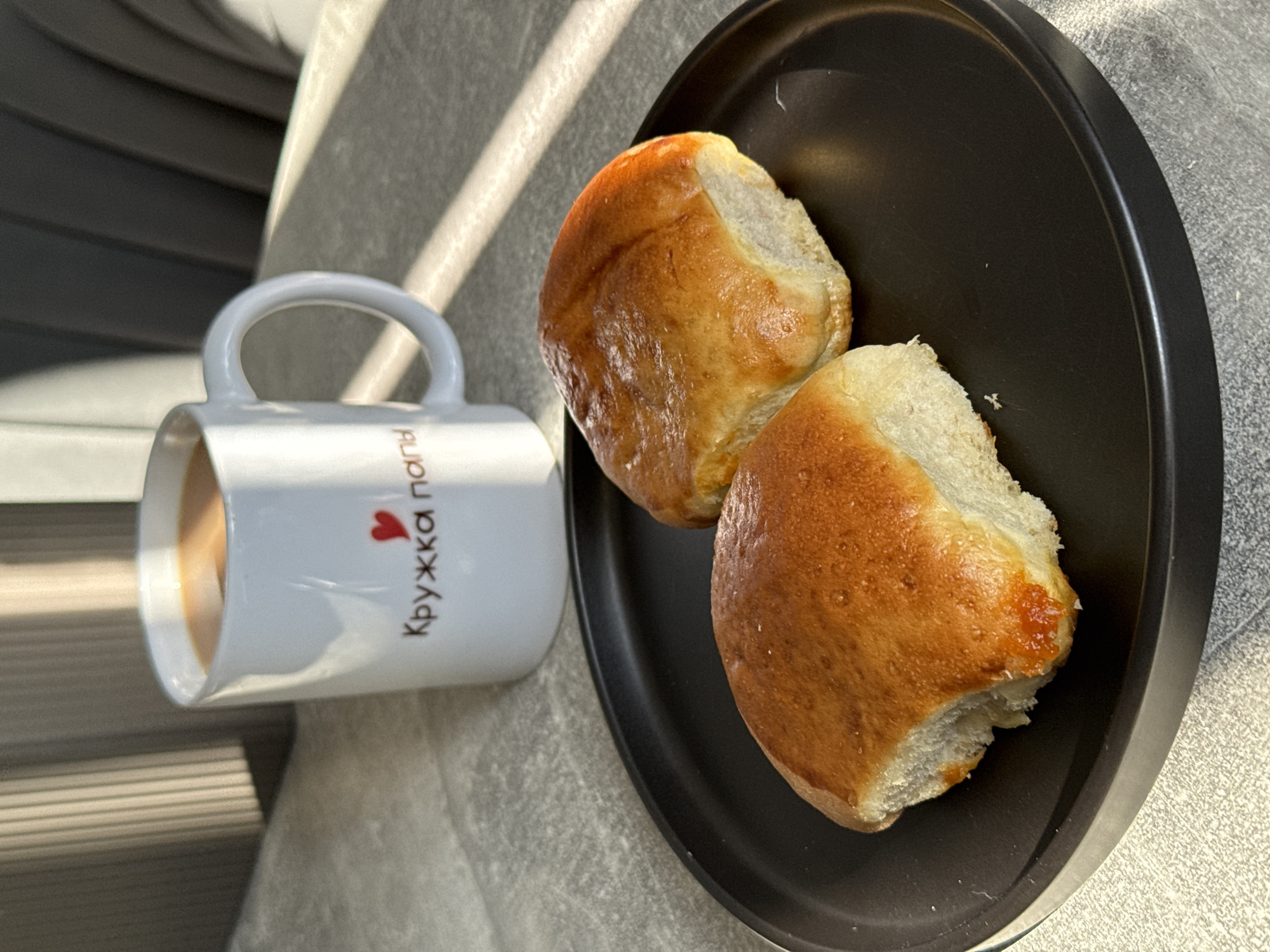 A white mug with "Кружка папы" (Dad's mug) written on it in red and a small heart is filled with a light brown liquid, likely coffee with milk. The mug is situated on a textured gray surface. In front of the mug, on a black circular plate, are two golden-brown baked goods. These appear to be sweet buns or pastries, with a slightly glossy top and a visible filling of a reddish-orange jam or preserves peeking out from one of them. The background shows vertical blinds casting striped shadows across the surfaces. The lighting suggests it is daytime, with sunlight creating highlights and shadows. The overall scene suggests a breakfast or snack setting, potentially in a home environment. No specific location in Lyubertsy, Russia is identifiable from the image.