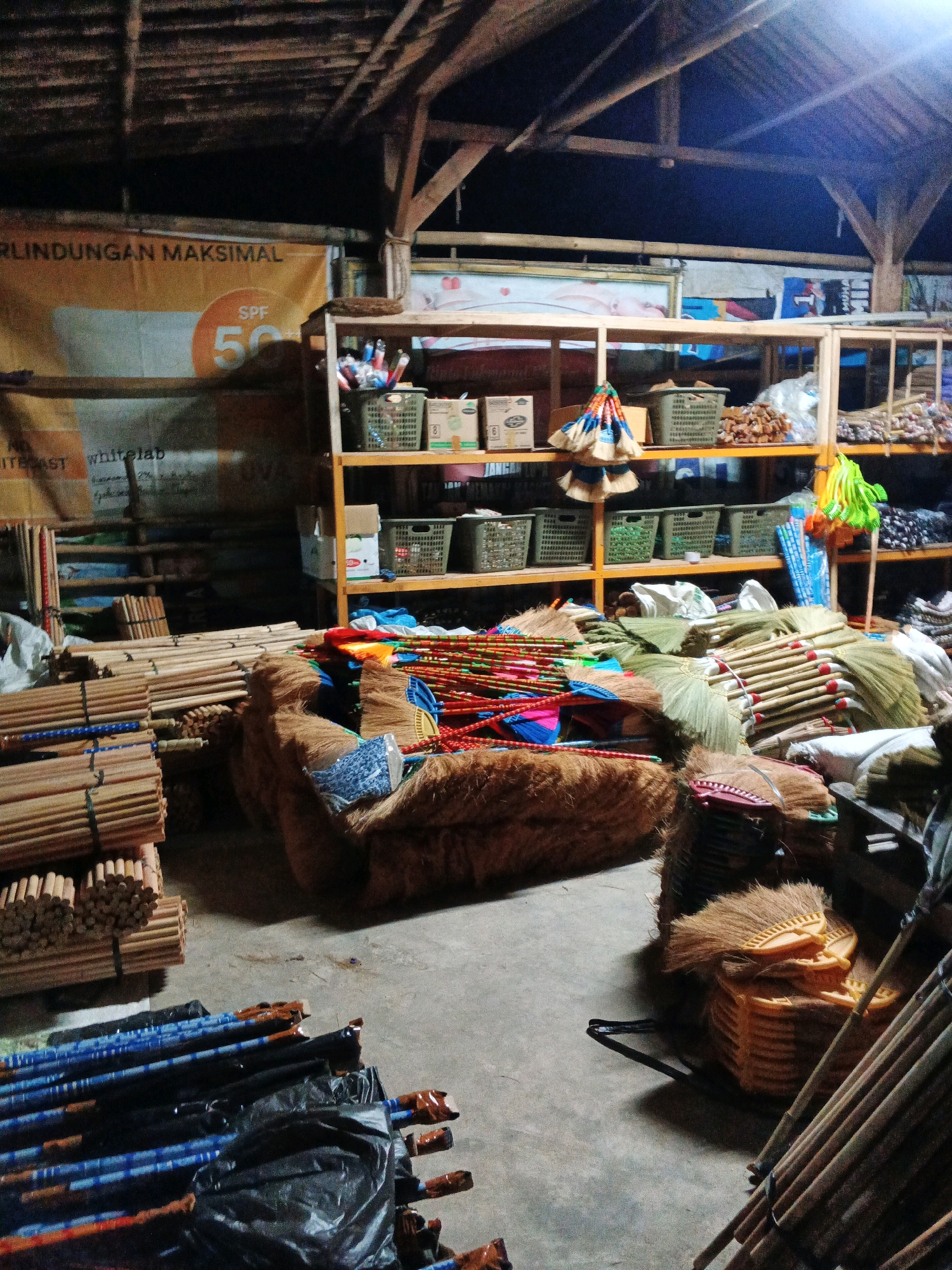 The image shows an indoor market stall in Sirau, Indonesia, filled with brooms and bamboo poles. The stall features wooden shelving units stocked with various items, including baskets, boxes, and more brooms. The floor is concrete and scattered with more brooms and bamboo poles. The ceiling is made of wood and bamboo. No people are visible in the scene.