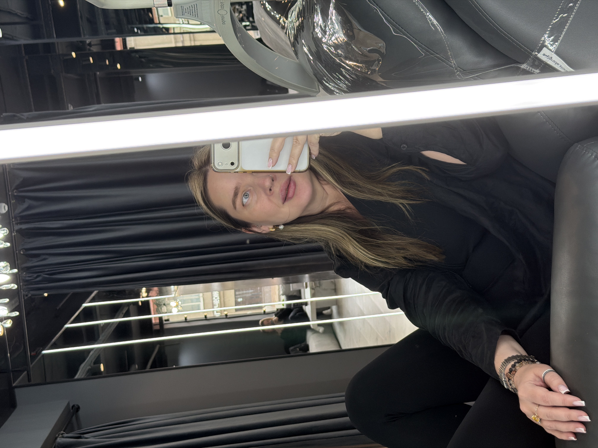 The image shows a woman taking a selfie in a mirror. She has blonde hair and is wearing a black shirt and black pants. She is seated in what appears to be a salon chair. Her reflection in the mirror shows a street scene in what is possibly Moscow, Russia. A building across the street is visible, and a person is walking on the sidewalk. The environment is modern and well-lit. The selfie is taken at an oblique angle, showing both the woman and her surroundings through the mirror.