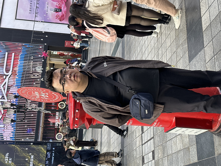 The image depicts a bustling pedestrian street scene in Yuzhong District, Chongqing, China. In the foreground, an adult male of East Asian descent stands facing the viewer, positioned to the left of a prominent red British-style pillar post box. He wears dark-framed glasses, a dark brown zip-up jacket over a black t-shirt, and dark pants, with a dark blue messenger bag slung across his body. His expression is neutral.
The red pillar post box, a distinct feature, has a circular red sign attached to its top, bearing white Chinese characters "重庆邮政" (Chongqing Post Office) and "CHONGQING POST OFFICE" in English.
In the midground and background, numerous other individuals populate the pedestrian area. To the left of the main subject, a child in a blue hooded jacket and light-colored pants walks away, looking left. Further right, a partial view of a person wearing black pants and sneakers is visible. To the right of the post box, several individuals stand, including a woman with long dark hair and a pink backpack, and another woman wearing a light-colored textured top, dark shorts, black sheer stockings, and a small brown and white shoulder bag.
The background features modern commercial buildings with large glass facades, displaying various retail signs. A prominent "MAC" logo is visible on one building. Other signs include "ONLY WEAR" and festive Chinese script, some indicating "Happy New Year." Decorative string lights with hanging bulbs are suspended across the street. The ground is paved with a pattern of grey and darker grey square and rectangular tiles.