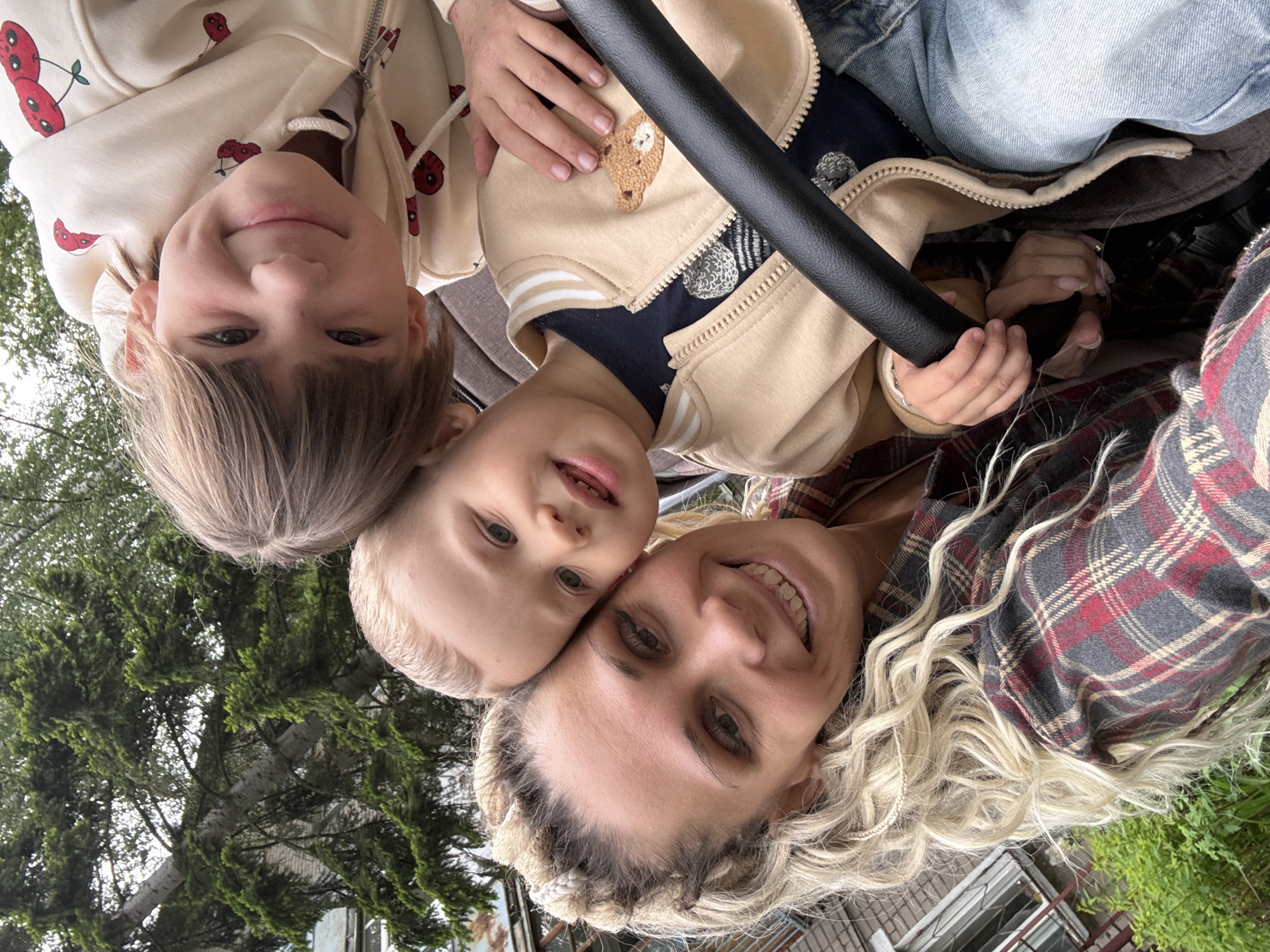 A close-up, slightly tilted shot shows a fair-skinned woman and three children, likely in Rusanovo, Russia. The woman, with blonde hair styled in braids, smiles broadly at the camera. A younger child with light blonde hair is nestled close to her, their heads touching. Above them, another child with dark blonde, fringe-cut hair, wears a hoodie with a cherry print. Next to the blonde-haired child is a child holding the handle of a baby stroller. The handle is black. The children wear clothing including a beige jacket with a bear patch and blue jeans. The woman wears a plaid flannel shirt. In the background, there is green foliage from trees and a partial view of a building. The scene appears to be outdoors, possibly in a park or residential area.
