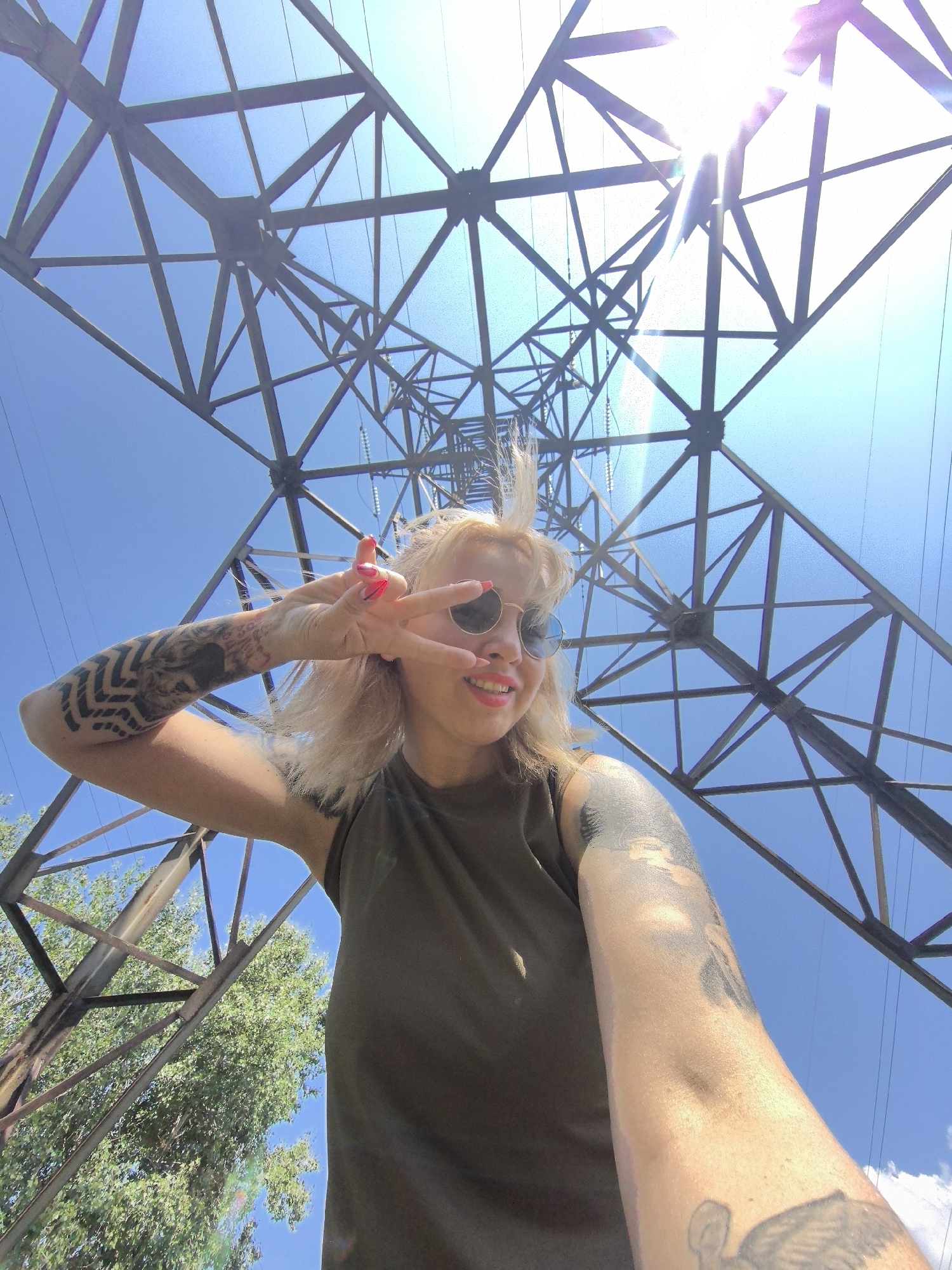 A young woman with blonde hair and sunglasses is taking a selfie outdoors. She is wearing a dark green t-shirt and has tattoos on her left arm and shoulder. Her left hand is raised, with her index and middle fingers extended in a peace sign. She is smiling and looking at the camera. The background consists of a large metal power line tower and a clear blue sky. The sun is visible in the upper right corner of the image, causing lens flare. Trees with green leaves are visible in the lower left corner. The woman's location is unknown, but it is not Noviy, Russia, as this city does not exist.