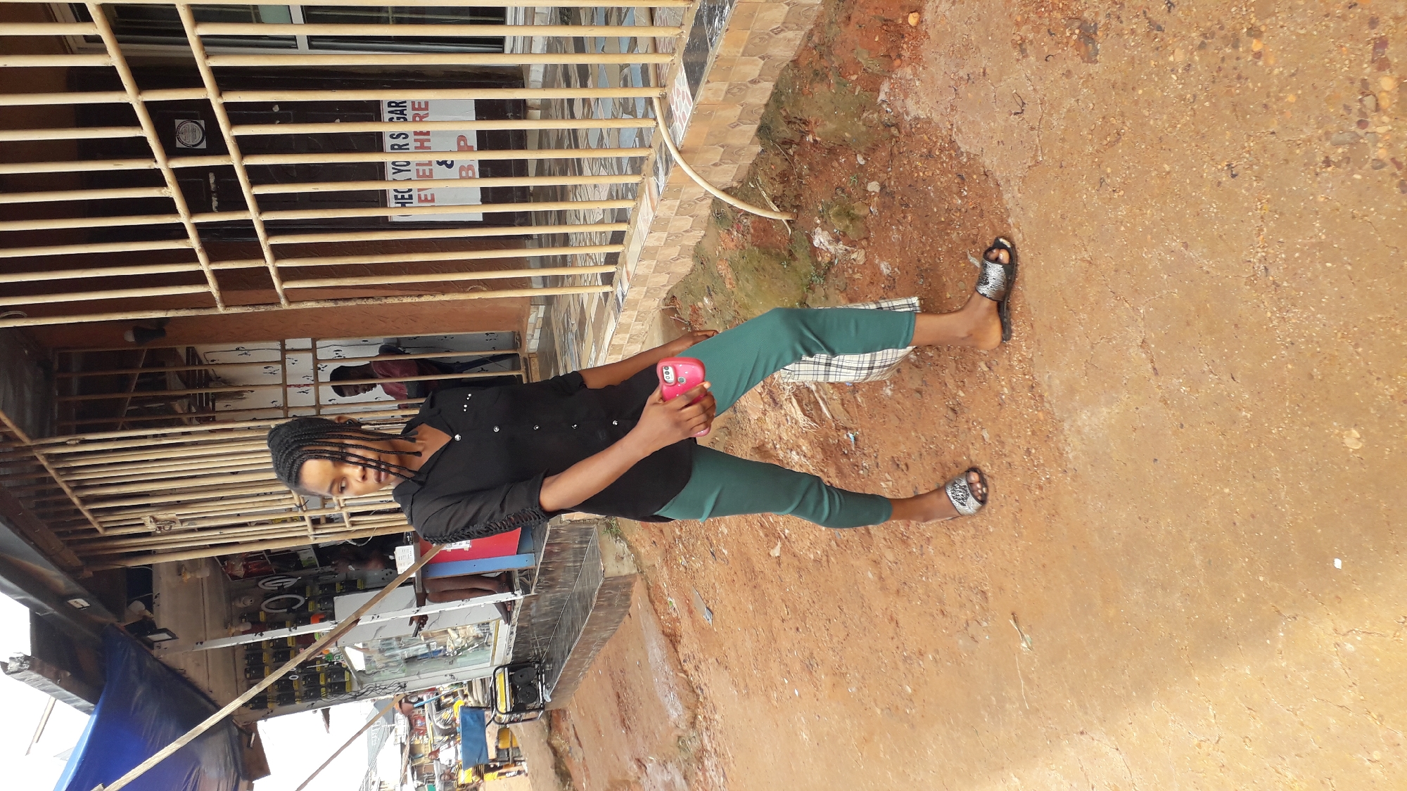A young woman with braided hair stands outside a shop in Nkpor, Nigeria. She is wearing a black button-down shirt and tight green pants, holding a pink smartphone in her right hand. A checkered bag rests on her left side, and she wears open-toed sandals on her feet. The shop behind her has a metal grate over its entrance, revealing shelves stocked with various items. The ground is covered in reddish-brown dirt.