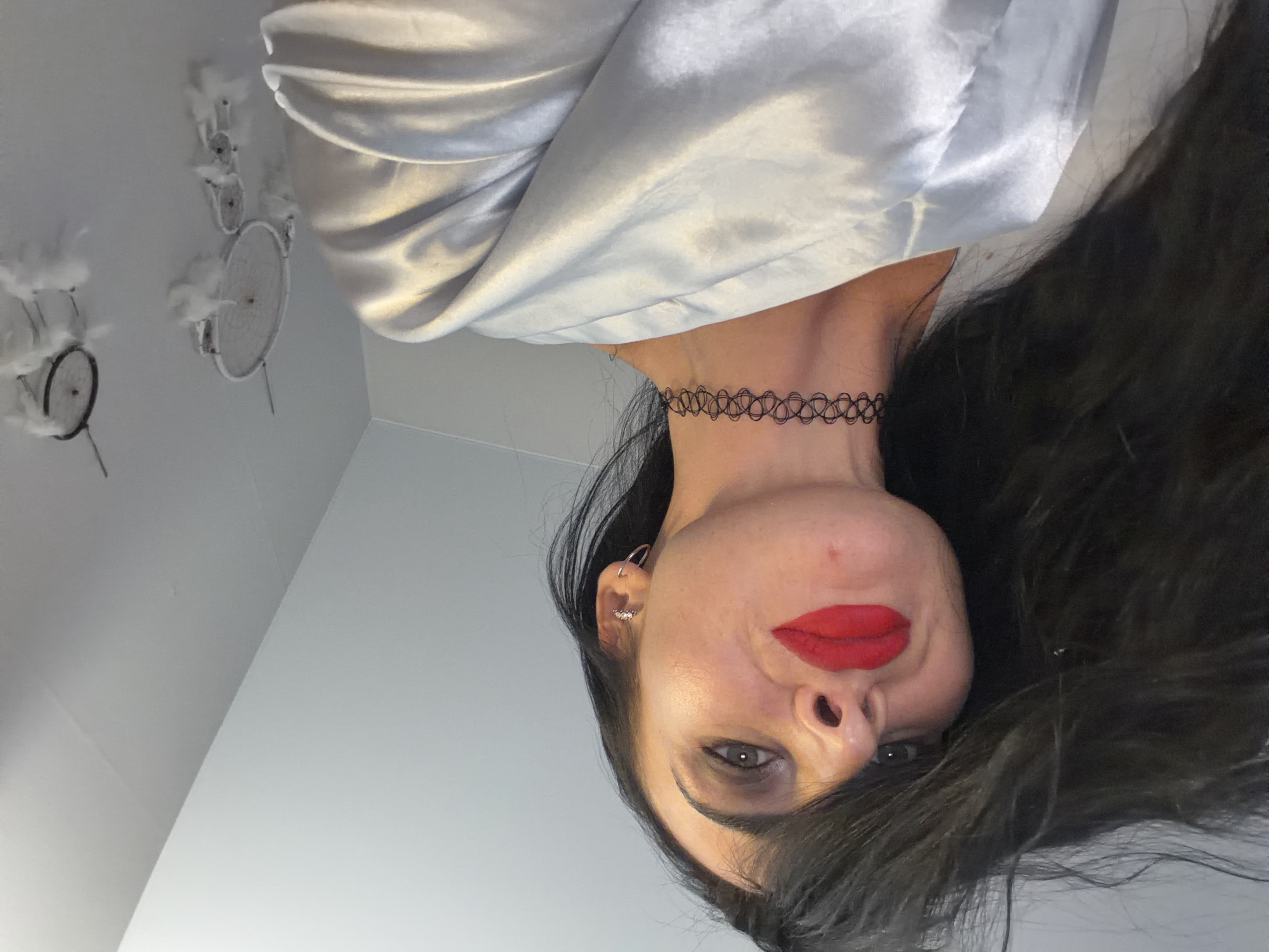 A medium-shot portrait depicts a woman with dark hair and fair skin. She wears a black, patterned choker necklace and red lipstick. She has multiple earrings in her left ear. She wears a shiny, light-colored garment that drapes around her shoulders. In the background, there is a white wall and ceiling. On the ceiling, several decorative objects are hung, appearing to be small, round frames with white feathers. The location is unknown.