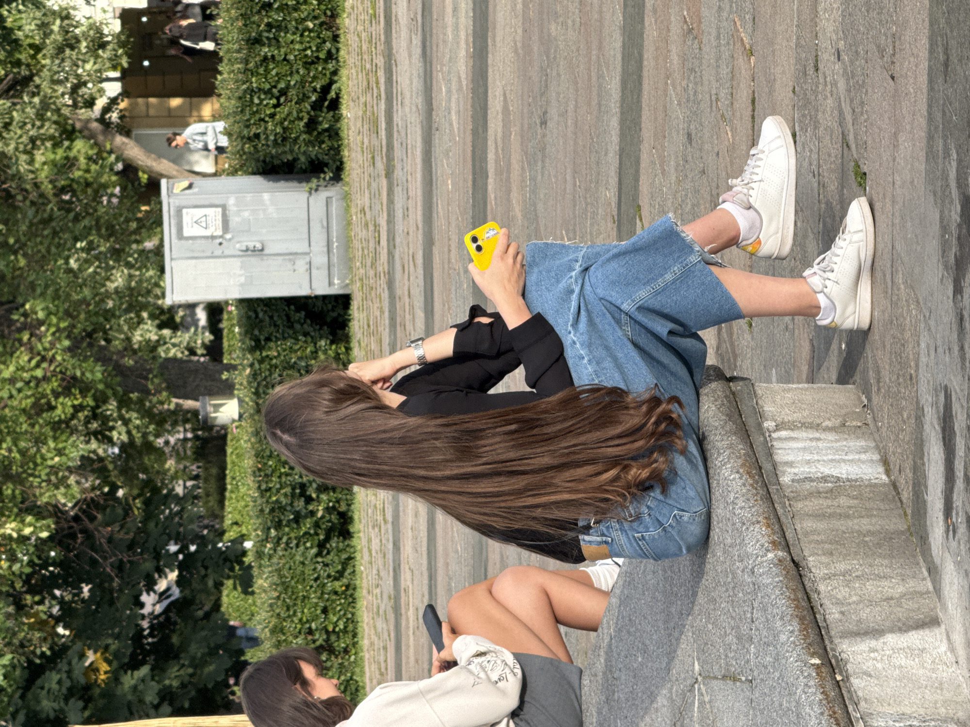 Two young women are sitting on a stone bench in an urban park. The woman on the left, with dark hair, is wearing a light-colored long-sleeved shirt and grey shorts. She is holding a black smartphone in her left hand. The woman on the right, with very long brown hair, is wearing a black long-sleeved shirt and wide-leg blue jeans. She is holding a bright yellow smartphone in her right hand. Both women are wearing white sneakers. In the background, there is a dense green hedge, a light grey electrical cabinet with a warning sign, trees, and other people walking in the distance. The ground is paved with stone tiles. This scene appears to be a public space within the city of Moscow, Russia.