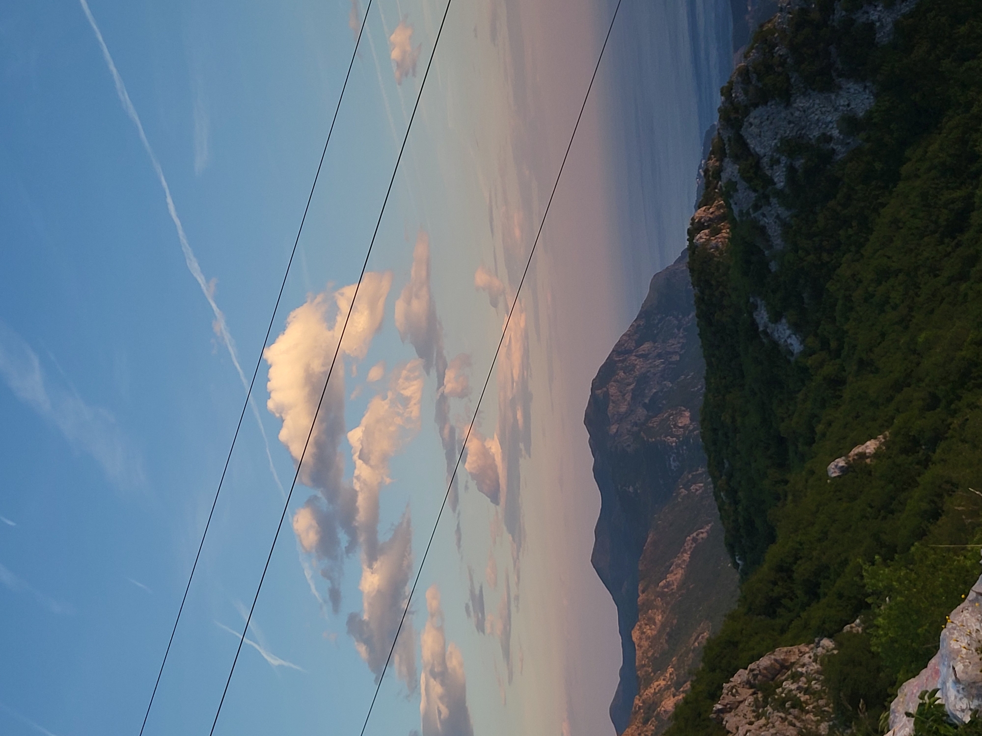 The image shows a view from Brajici, Montenegro, featuring a landscape of mountains, sea, sky, and clouds. Several power lines cut diagonally across the scene from the upper right to the lower left. In the sky, there are contrails, indicating jet travel. The mountains are covered in vegetation and rocky terrain. The sea is a deep blue, contrasting with the pastel colors of the sky. The sky is predominantly blue with patches of white and pink clouds, suggesting sunrise or sunset.