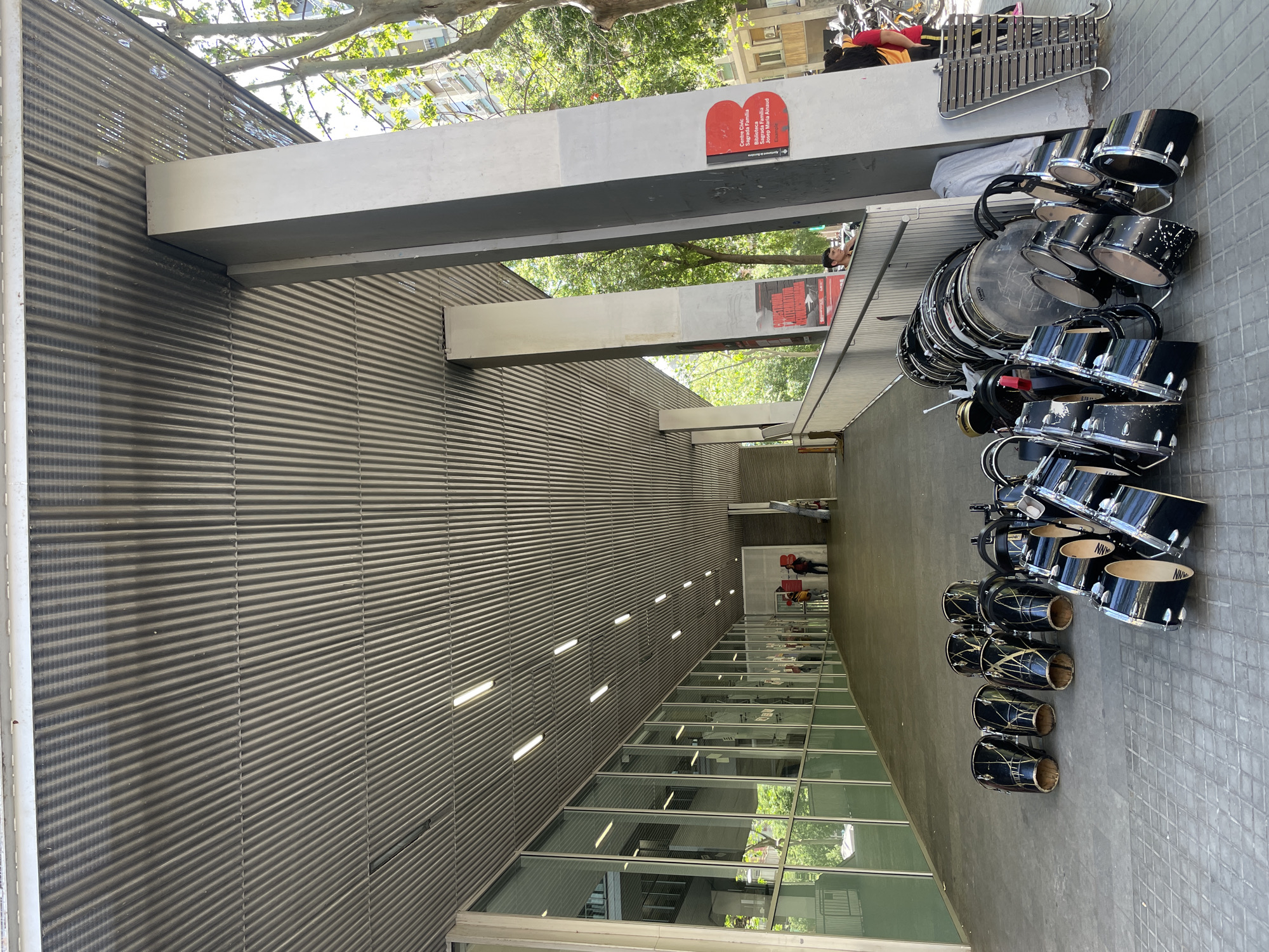 The image shows a collection of drums and percussion instruments assembled on a paved surface next to a building. The building has a corrugated metal facade, a row of windows, and overhead beams. A sign on one of the beams reads "Centre Civic Sagrada Familia Josep Maria Pemanrd." In the background, a person can be seen walking away from the drums and into the building. There is a xylophone to the right of the drums. The location is Barcelona, Spain, given the name of the Sagrada Familia on the sign.