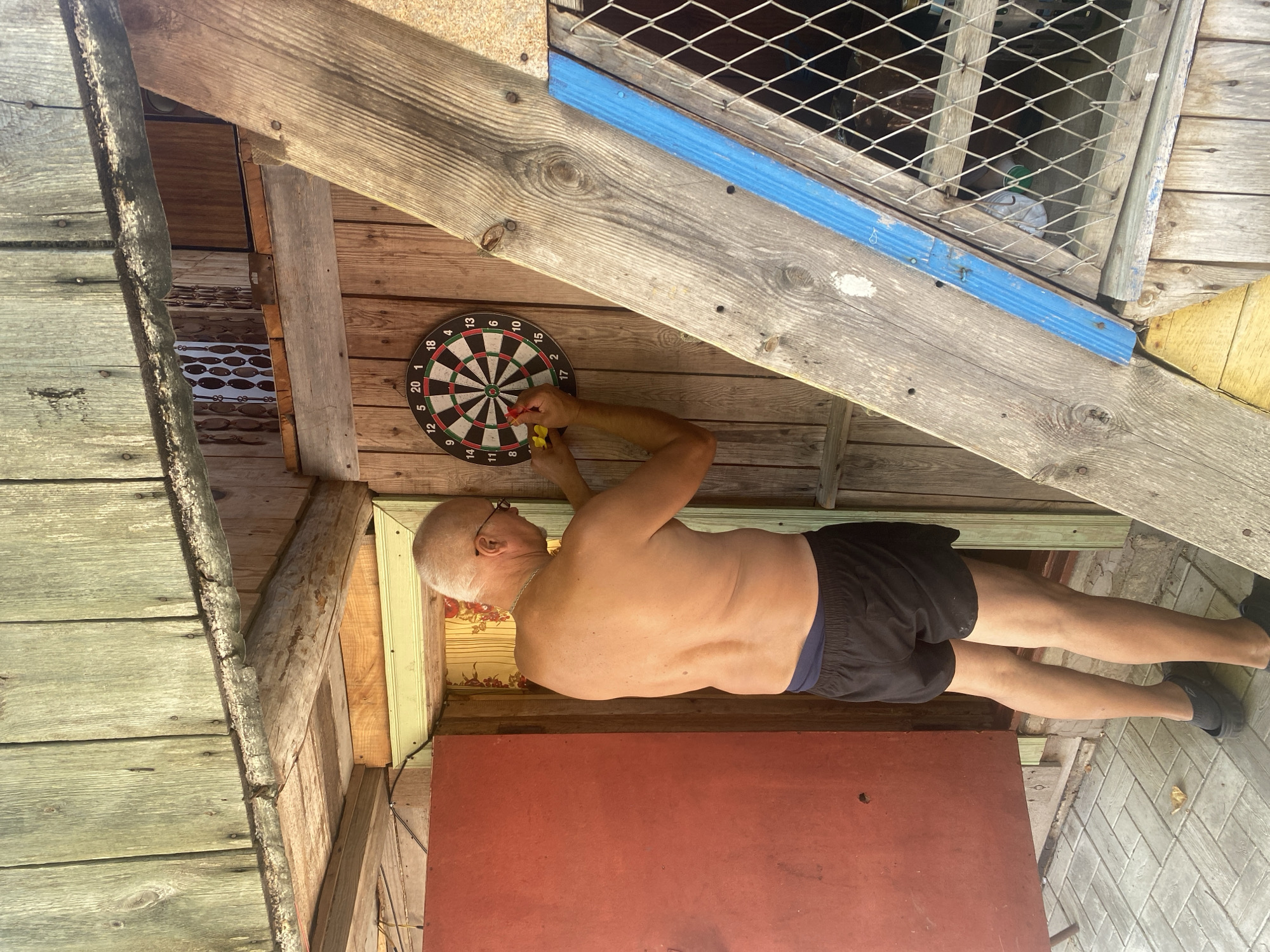 An adult Caucasian male, with short grey hair and glasses, is standing outdoors in front of a wooden shed. He is shirtless and wearing black shorts and black clogs. He is holding two darts and is about to throw one at a dartboard mounted on the shed wall. The dartboard is a standard circular design with numbers. The shed appears rustic, with weathered wooden planks. A red door is visible to the left of the shed, and a window with wire mesh is to the right. The ground is paved with grey tiles. The overall setting suggests a casual, outdoor recreation area. The location is Krupki, Belarus.