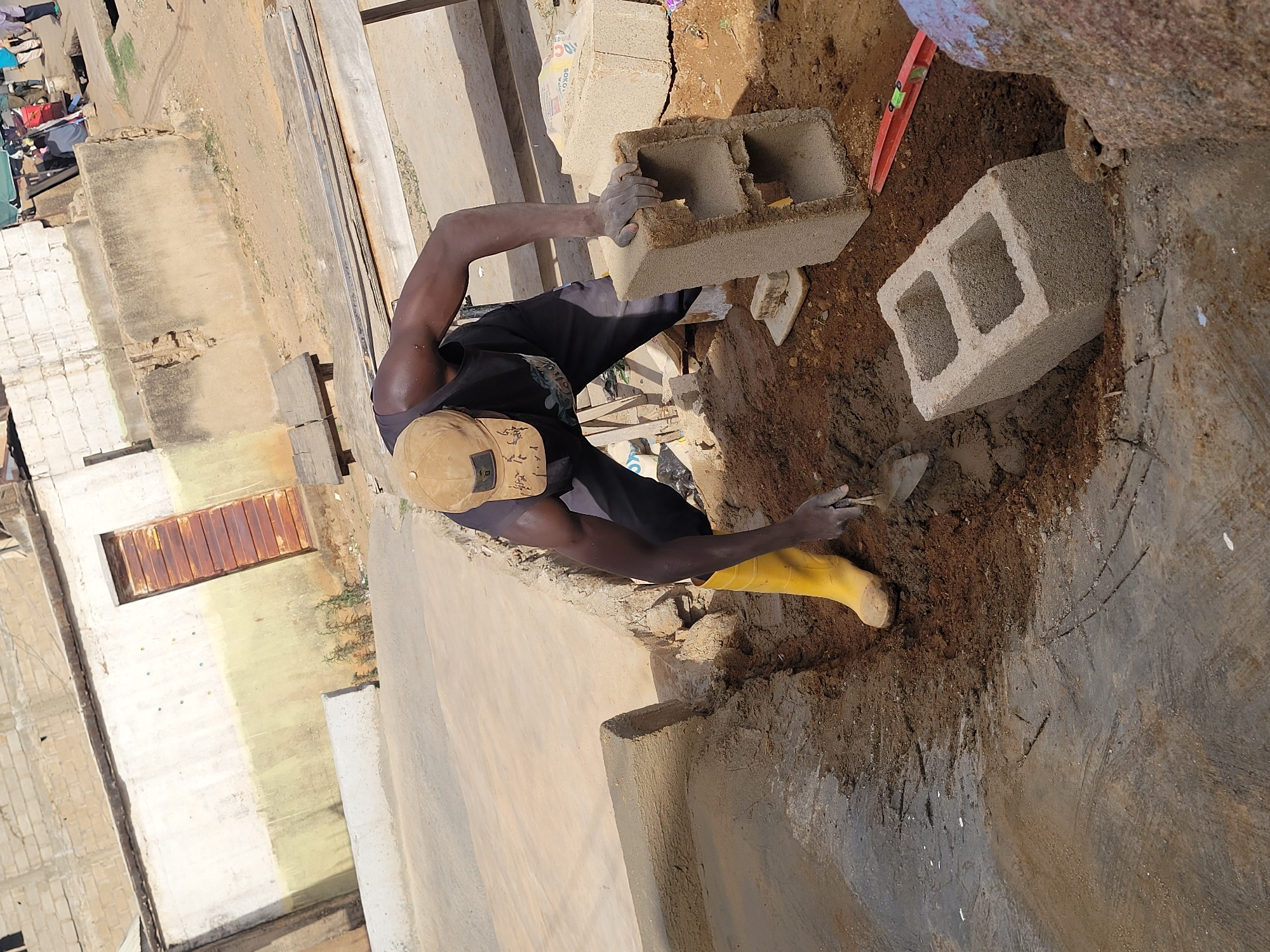 This overhead shot captures a man actively engaged in construction work at an outdoor site, likely in Jos, Nigeria, given the provided context. The image is rotated, showing the ground plane vertically.
The central subject is a dark-skinned man, seen from above, wearing a black t-shirt, dark trousers, a tan baseball cap with some illegible writing, and distinctive bright yellow boots. His left arm shows visible musculature. He is in the process of laying concrete blocks, holding a hollow block with both hands, which are covered in mortar or dirt. Another similar hollow block is visible on the ground nearby, along with a red and green spirit level.
The setting is a rugged construction area with loose brown soil and partially constructed concrete elements, including a newly laid section of concrete or mortar in the foreground. In the background, there are other buildings under construction or in disrepair, made of light-colored blocks or concrete, and what appears to be laundry hanging to dry. A partially visible sack in one of the cropped images shows the text "SOFO CEMENT". Wooden planks are also scattered around the site.
The scene appears to be taking place during the daytime under bright sunlight, indicated by the clear lighting and shadows. The man appears focused on his task, contributing to the development of the structure.