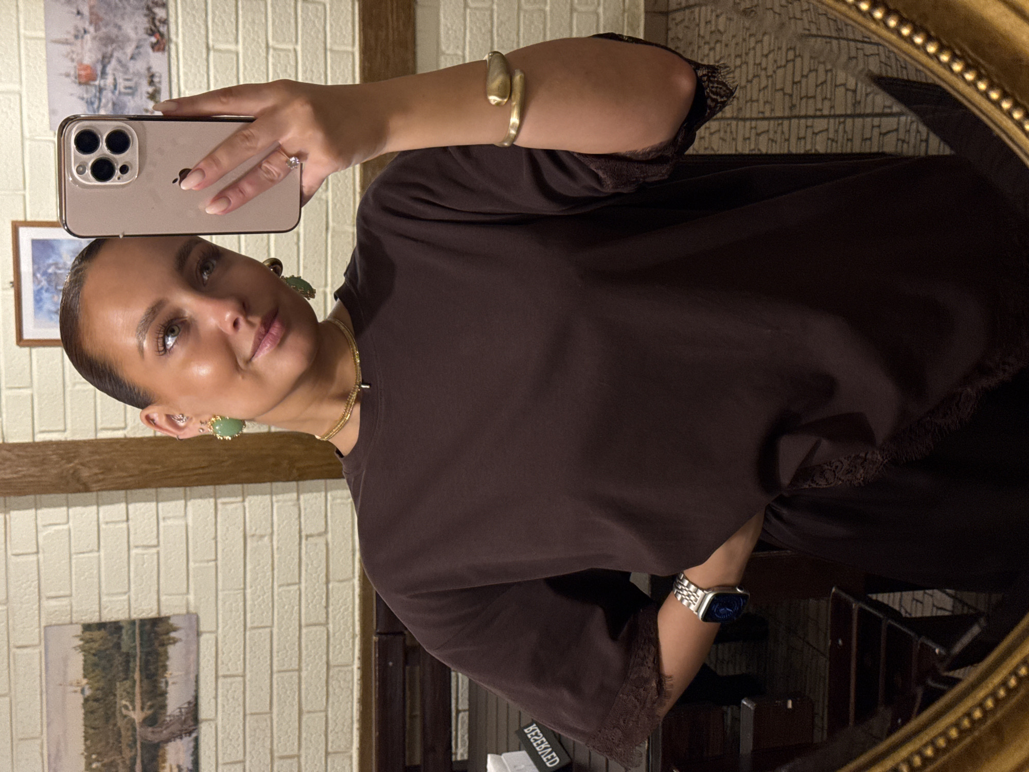 The image shows a woman taking a selfie in front of a mirror. She is wearing a brown top with lace details, gold jewelry (earrings, necklace, bracelet, and a watch on her wrist), and holding an iPhone. The background includes a brick wall, framed pictures, and what appears to be the interior of a room. It's not possible to confirm the location in Chaikovsky, Russia from the image alone.