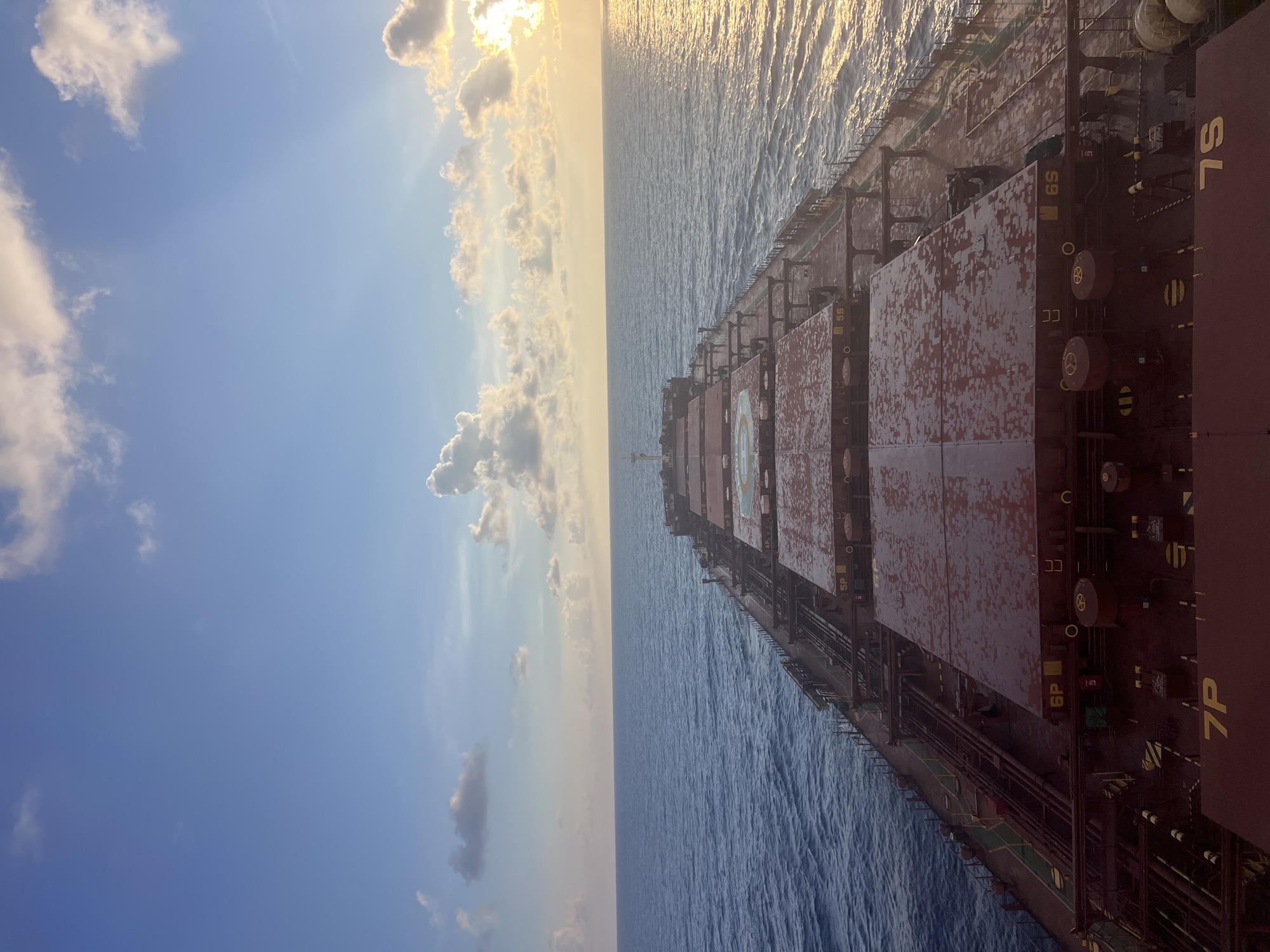 The image is a high-angle shot of a large cargo ship at sea. The ship is the primary subject, with its long, red deck stretching towards the horizon. Several large, hatch covers are visible, marked with numbers such as "7P," "6P," and "6S." The sea is a deep blue, with gentle waves reflecting the sky. The sky is a mixture of blue and white clouds, with the sun setting on the right side of the frame, casting a warm, golden light across the water and sky. The overall impression is one of vastness and solitude, with the ship traversing a calm sea under a dynamic sky.
