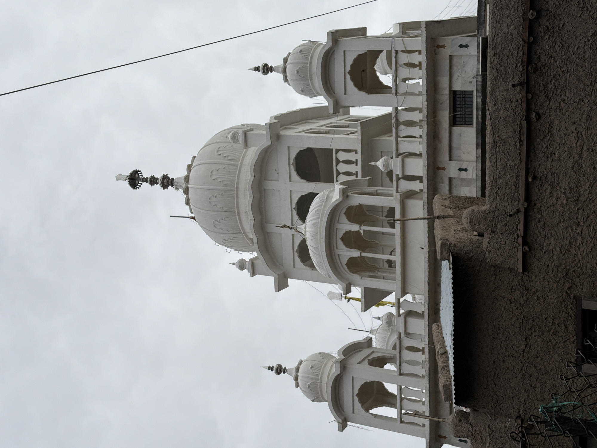 This is a low-angle, eye-level, exterior, full shot of a white Sikh Gurdwara in Leh, India. The Gurdwara has multiple domes and ornate white stonework. The sky is overcast and gray. In the foreground, there is a rough, textured mud-and-stone wall topped with a corrugated metal roof. A dark wire runs diagonally across the upper right portion of the frame. A chain-link fence is partially visible in the lower left corner. There are no people or animals in the image.