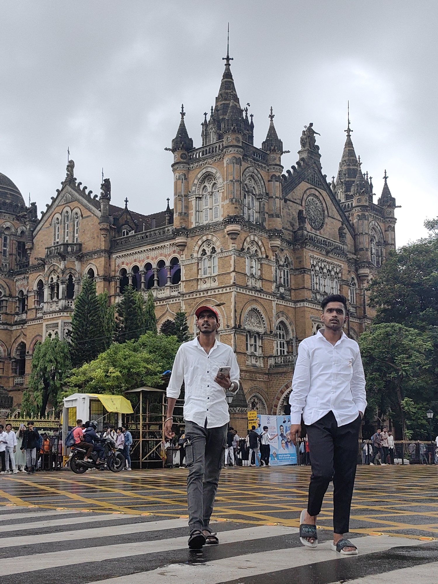 Two men, one wearing a red cap and a white shirt with dark grey pants, and the other wearing a white shirt and black pants, walk across a wet pedestrian crossing. They are in front of the Chhatrapati Shivaji Maharaj Terminus, a historic railway station in Mumbai, India. The building, characterized by its Victorian Gothic Revival architecture, features intricate stonework, spires, and large windows. The sky is overcast, and trees are visible behind the building and in the background. Several other people and a motorcycle are also present in the scene, with some gathered near a small kiosk.