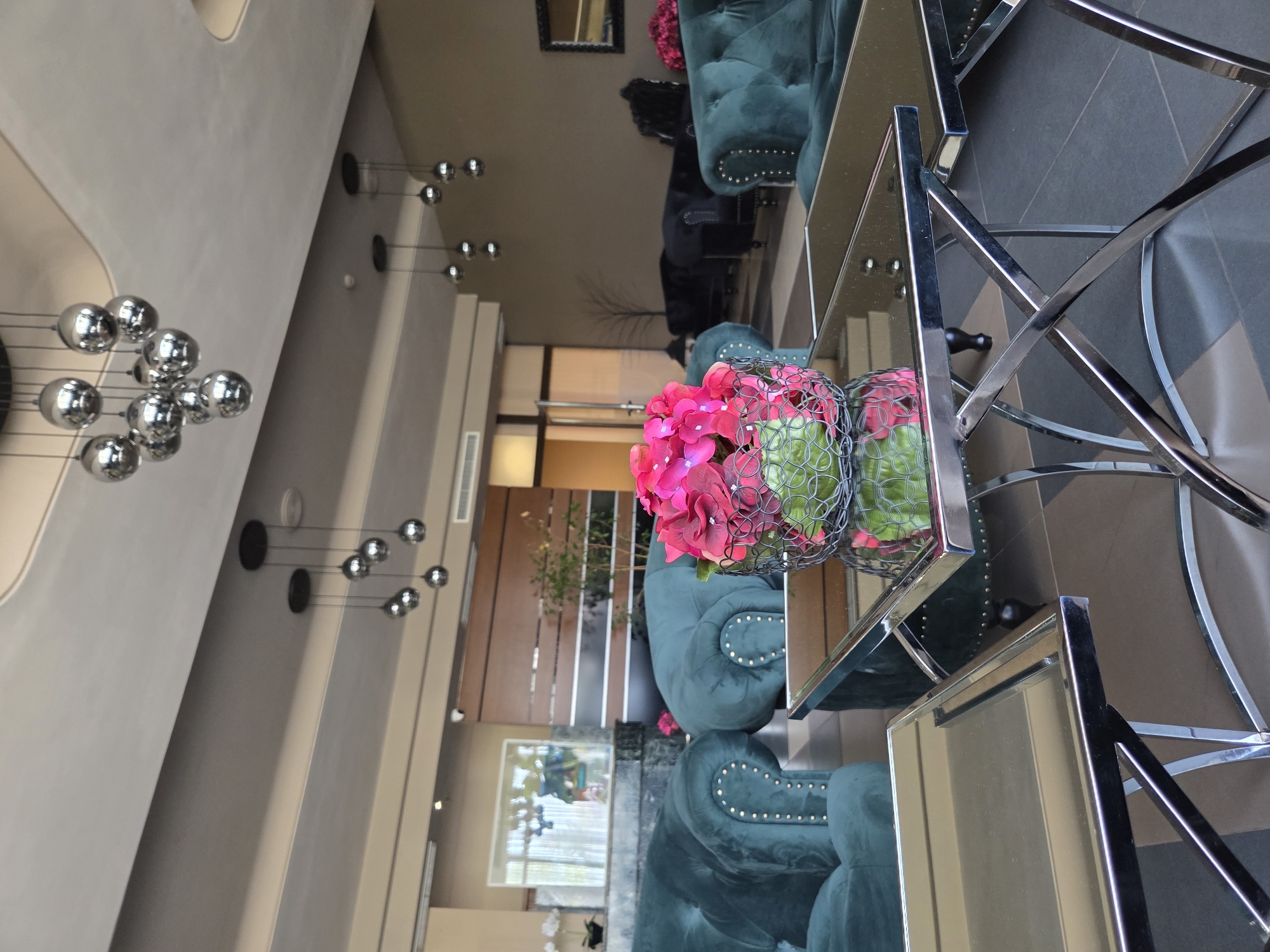 The image shows the interior of a building, possibly a hotel or lobby, in Pleven, Bulgaria. The main focus is on a modern table with a reflective surface, on which there is a floral arrangement of pink flowers in a wire basket. The table has a chrome frame with an intricate design.
Behind the table are plush, dark teal-colored sofas or chairs, adorned with silver studs. In the background, there is a glimpse of a lobby area with various seating arrangements, some of which are dark or black. A series of modern, spherical pendant lights hang from the ceiling, contributing to the contemporary design of the space.
There is no indication of people or specific actions in the immediate foreground of the image. The setting appears to be a designed interior space, likely for receiving guests or providing a comfortable waiting area. The image suggests a sense of luxury and modern design.