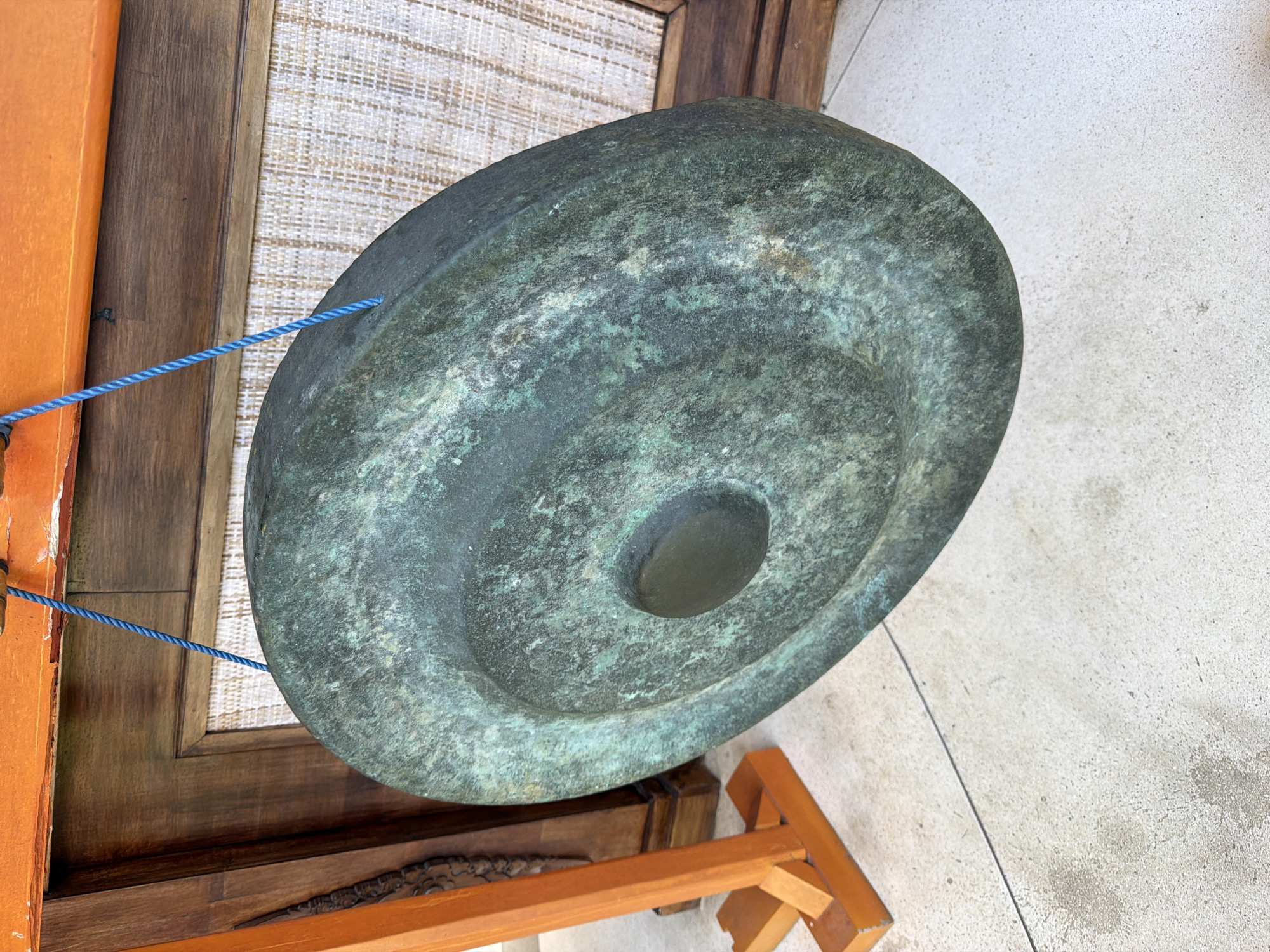 The image shows a large, round, greenish-blue metal gong, likely made of bronze due to the color. It is mounted on a wooden stand. The gong has concentric circles pressed into its surface. A blue rope is attached to the gong's frame. The scene appears to be outdoors, possibly on a tiled patio. Given the location information, the gong is likely located in Peliatan, Indonesia.