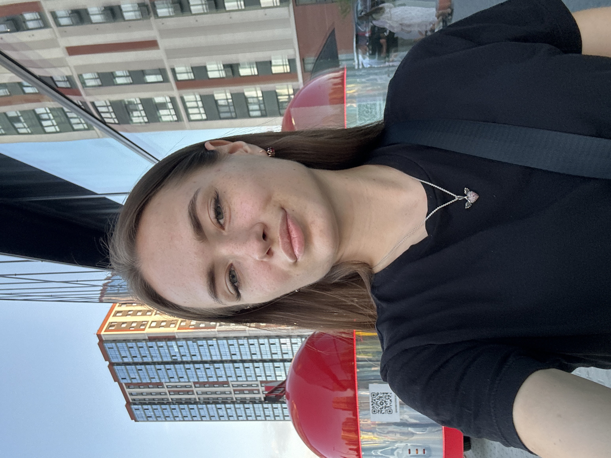 The image features a young woman taking a selfie. She is wearing a black t-shirt and a necklace with a small heart-shaped pendant. Her head is tilted, and she is looking towards the camera with a slight smile. The background shows a reflection of modern buildings with many windows, suggesting an urban environment. The sky is clear, indicating it's daytime. Part of a red object, possibly a bus or a kiosk, is visible in the foreground. There is a QR code on the red object. The image appears to be taken from a slightly low angle.