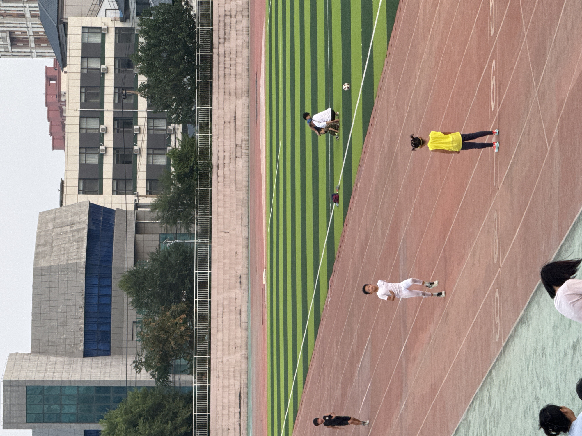 The image depicts a wide shot of a sports stadium with a track and field area. In the foreground, on the track, a young boy in a white t-shirt and white pants is running. Further down the track, another young boy in a black tank top and black shorts is also running. On the green and black striped field, two figures are interacting, one in a white t-shirt and the other in a camouflage-patterned outfit; a soccer ball is nearby. In the far right foreground, a young girl in a yellow vest and dark pants stands on the track, facing away from the camera. In the background, there are several buildings, including a modern concrete building with blue glass windows on the left and a multi-story building with many windows and air conditioning units on the right. Trees are visible between the buildings and the stadium. The sky is overcast. The setting is identified as Linghe, China.