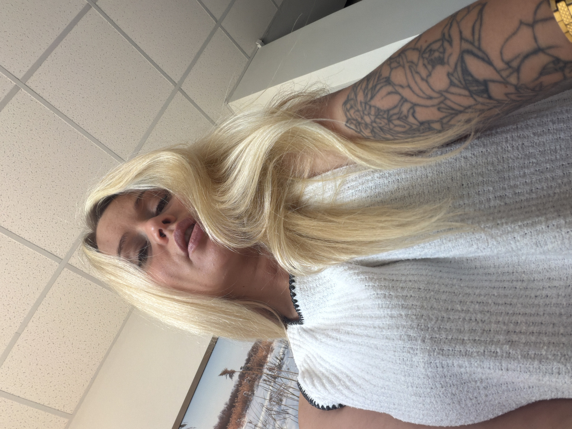 A woman with blonde hair is looking down at the camera. She is wearing a white top with black trim. She has a tattoo of flowers on her arm. She is in an indoor setting with a drop ceiling. A framed picture of a winter landscape is visible in the background. The image was taken in Sarapul, Russia.