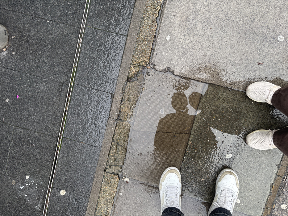An overhead view depicts two individuals standing on a wet, tiled pavement in an urban environment. The ground consists of various grey surfaces: dark grey square tiles, a lighter grey concrete section, and a rougher, multi-toned stone curb separating them. Puddles of water, reflecting ambient light, are present on the darker tiled areas, particularly beneath and between the individuals' feet. Scattered white specks, consistent with discarded chewing gum, and a small pink fragment are visible on the tiles. A narrow, parallel metallic strip, possibly a drainage channel or decorative inlay containing minimal green debris, runs horizontally across the upper third of the image within the dark grey tiles. The individual on the left wears white sneakers with beige soles and dark trousers. The individual at the bottom right wears white mesh sneakers and brown trousers. The scene indicates recent rain or damp conditions. Based on the request, this scene is located in the city of London, United Kingdom, though specific identifying landmarks within the image itself are absent.