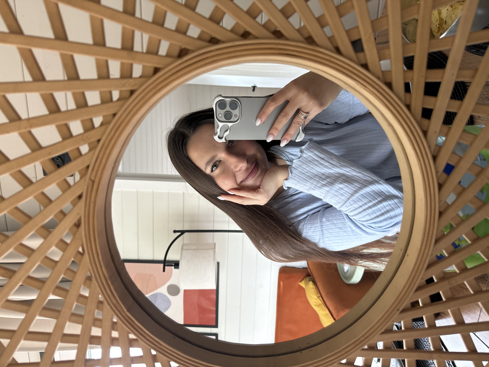 A woman is taking a selfie in a round mirror with a wooden lattice frame. She is wearing a light blue shirt and holding a grey smartphone with a distinctive case. Her long, brown hair is visible. The background, seen through the mirror, includes a white wall with vertical planks, a painting with geometric shapes, a floor lamp, and a glimpse of an orange sofa with a yellow accent. The reflection also reveals other objects on a table. The location is Литвиново, Russia.