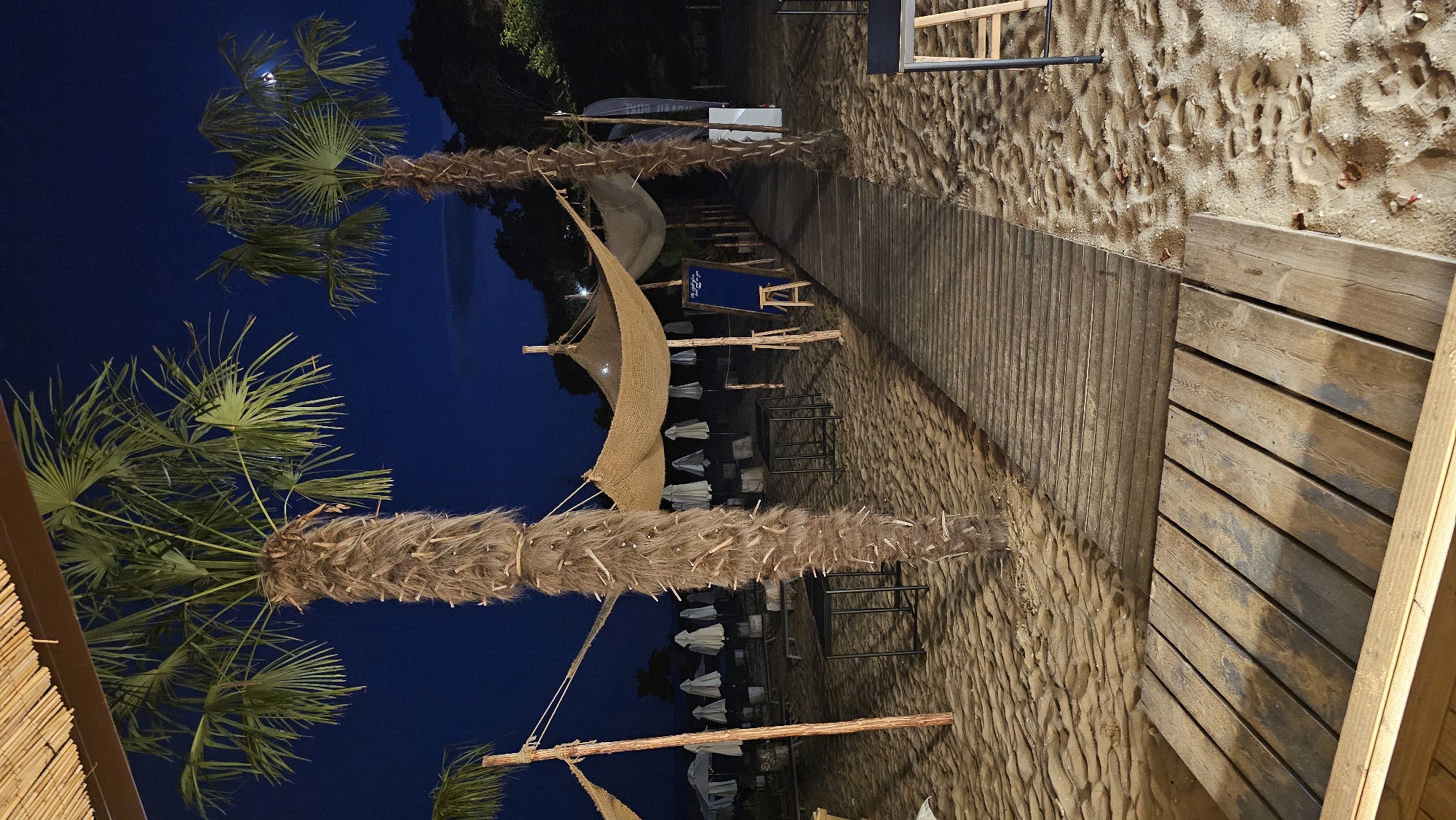 The image shows a beach at night, likely in Varna, Bulgaria. A wooden boardwalk runs through the sand, leading towards a shaded area with tables and chairs. Palm trees are visible, and beach umbrellas are set up in the background. The sky is dark blue, indicating it is nighttime. There are no people visible in the image.