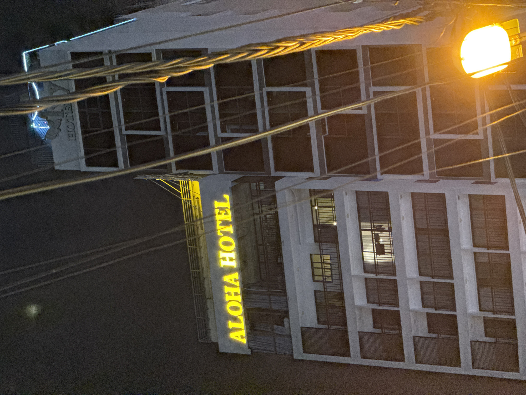 The image shows the Aloha Hotel in Nha Trang, Vietnam, at night. The hotel is a multi-story building with a white facade and balconies. The "ALOHA HOTEL" sign is illuminated in yellow neon. Power lines run across the foreground, partially obscuring the view of the building. There is a bright streetlight in the bottom right corner of the image. The sky is dark, with a faint, blurred object that may be the moon or a light in the distance. The windows of the hotel are dark, with some showing interior lights. The overall scene suggests a typical urban night view.