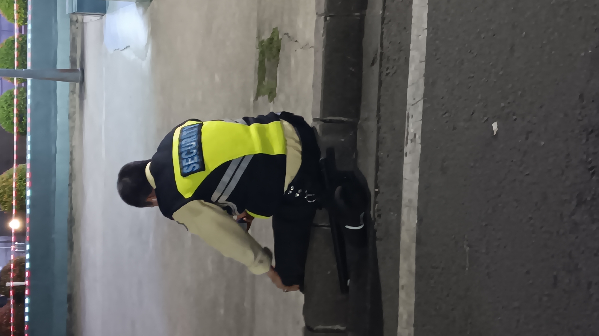A security guard in a yellow and navy uniform with "SECURITY" printed on the back is sitting on a concrete curb at night in South Jakarta, Indonesia. The guard is holding a smartphone and appears to be looking at it. A dark baton is lying on the ground in front of the guard. The background shows a dimly lit street with a white line marking a crosswalk and some decorative plants and lights in the distance.