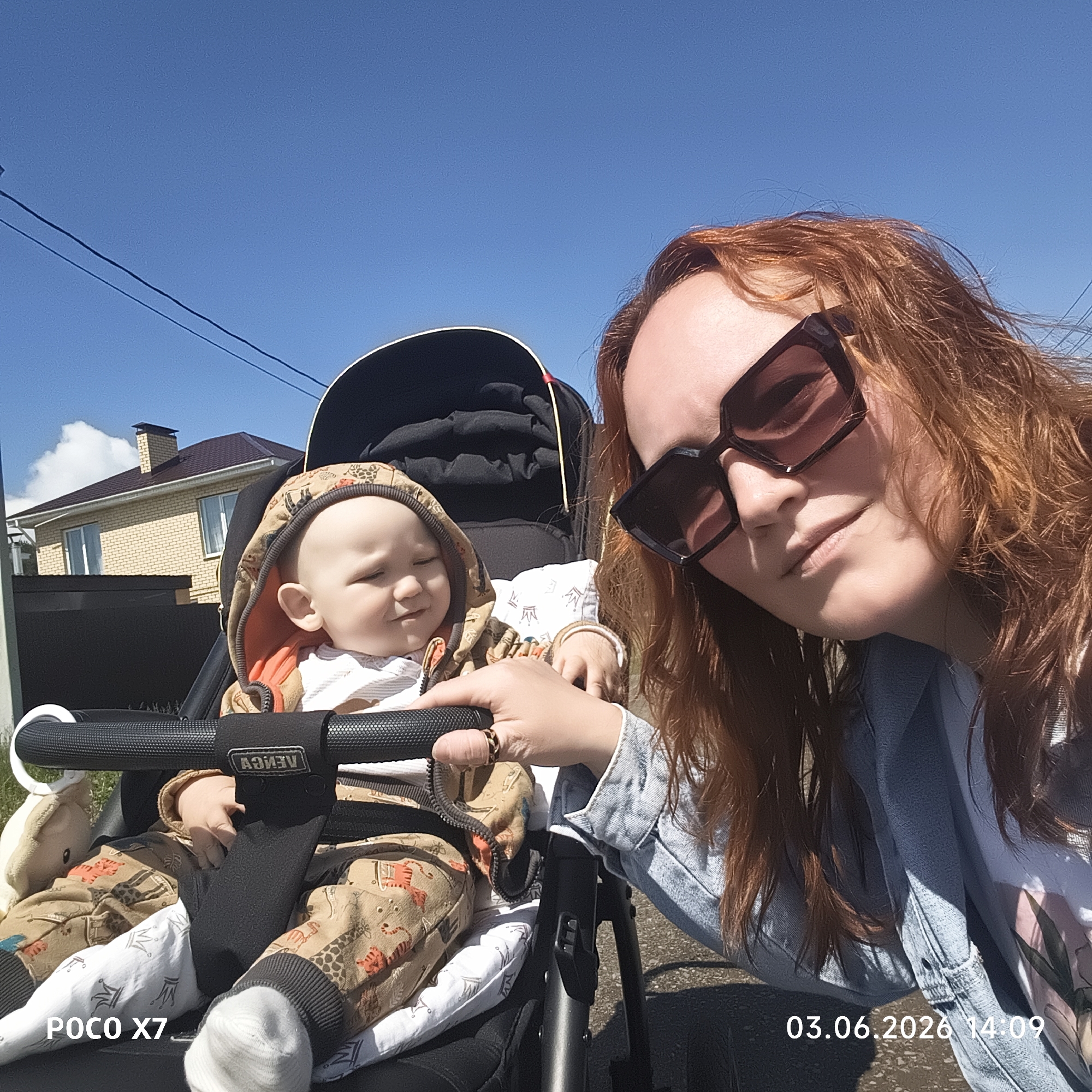 This sunny outdoor selfie captures a woman and a baby in a stroller, likely during an outing in Chaikovsky, Russia. The woman, positioned closer to the camera, has long reddish-brown hair, wears large dark sunglasses, and a light blue denim jacket, exhibiting a slight smile. Her right hand rests on the black handlebar of the stroller.
Seated in the dark-colored stroller is a baby, appearing to be an infant or toddler. The baby is dressed in a hooded light-colored outfit adorned with brown animal patterns, including giraffes and tigers, and white socks. The baby's eyes are squinted, possibly from the bright sunlight. A white plush toy hangs from the stroller's handlebar, which also displays the brand name "VENGA" on a central strap.
The background reveals a clear, vibrant blue sky with a few small white clouds. On the left side, a light-colored brick house with a dark roof and chimney is visible, suggesting a residential setting. Power lines traverse the upper left portion of the image.
The photo is brightly lit, indicative of a clear, sunny day. Visible text includes "POCO X7" in the bottom left corner, identifying the camera device, and "03.06.2026 14:09" in the bottom right, indicating the date and time the photograph was taken.