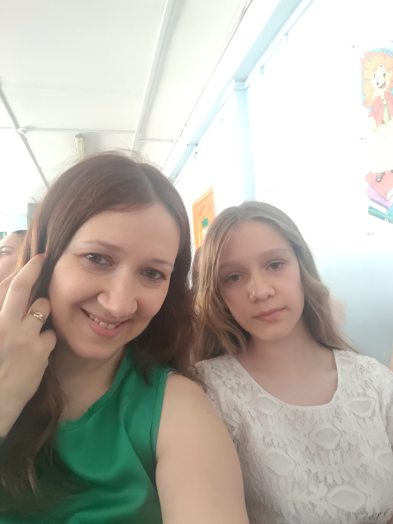 The media file is a close-up selfie featuring two people. On the left is an adult woman with brown hair and a green sleeveless top. She is smiling and touching her hair with her right hand, which has a ring on her finger. To her right is a younger girl with long blonde hair and a white lace top. She is looking directly at the camera with a neutral expression.
The setting appears to be indoors, possibly a classroom or a common area, given the ceiling lights and what looks like a decorated wall in the background. On the right wall, there is a colorful drawing of a cartoon lion standing on books, and a stack of books. The lighting suggests it is daytime. The overall mood is casual and friendly. There is no visible text in the media file. The location context provided is Rostov-on-Don, Russia.