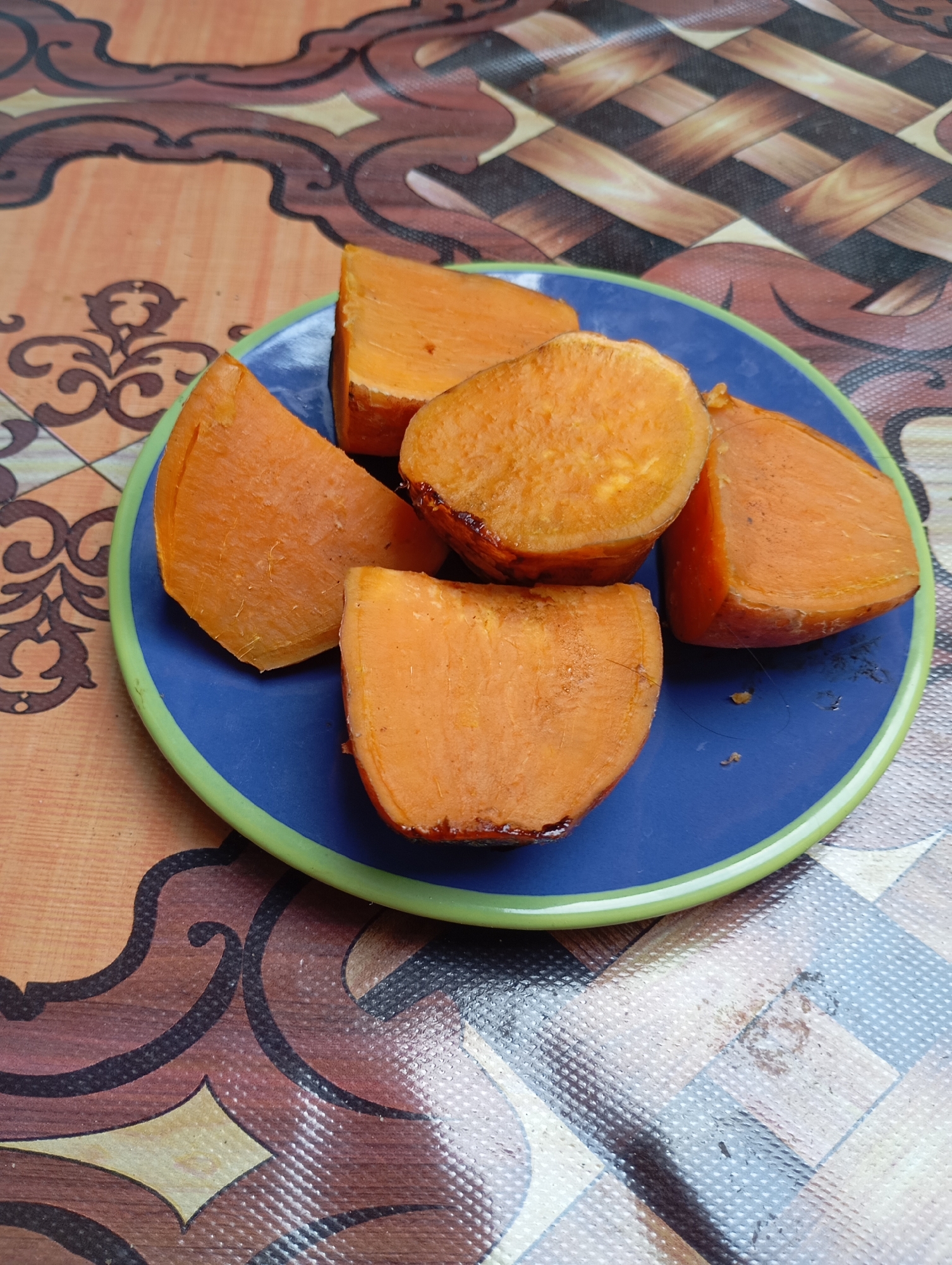 A blue plate with a green rim contains several wedges of baked sweet potato. The plate rests on a patterned tablecloth. The tablecloth has a design of interlocking geometric shapes and floral motifs in brown, orange, and black. The sweet potato pieces are a vibrant orange color, with some exhibiting a slightly caramelized or browned exterior. The scene is captured from a slightly elevated, close-up perspective. The image does not provide specific information about the location within Balikpapan, Indonesia, beyond being a domestic or food-related setting.