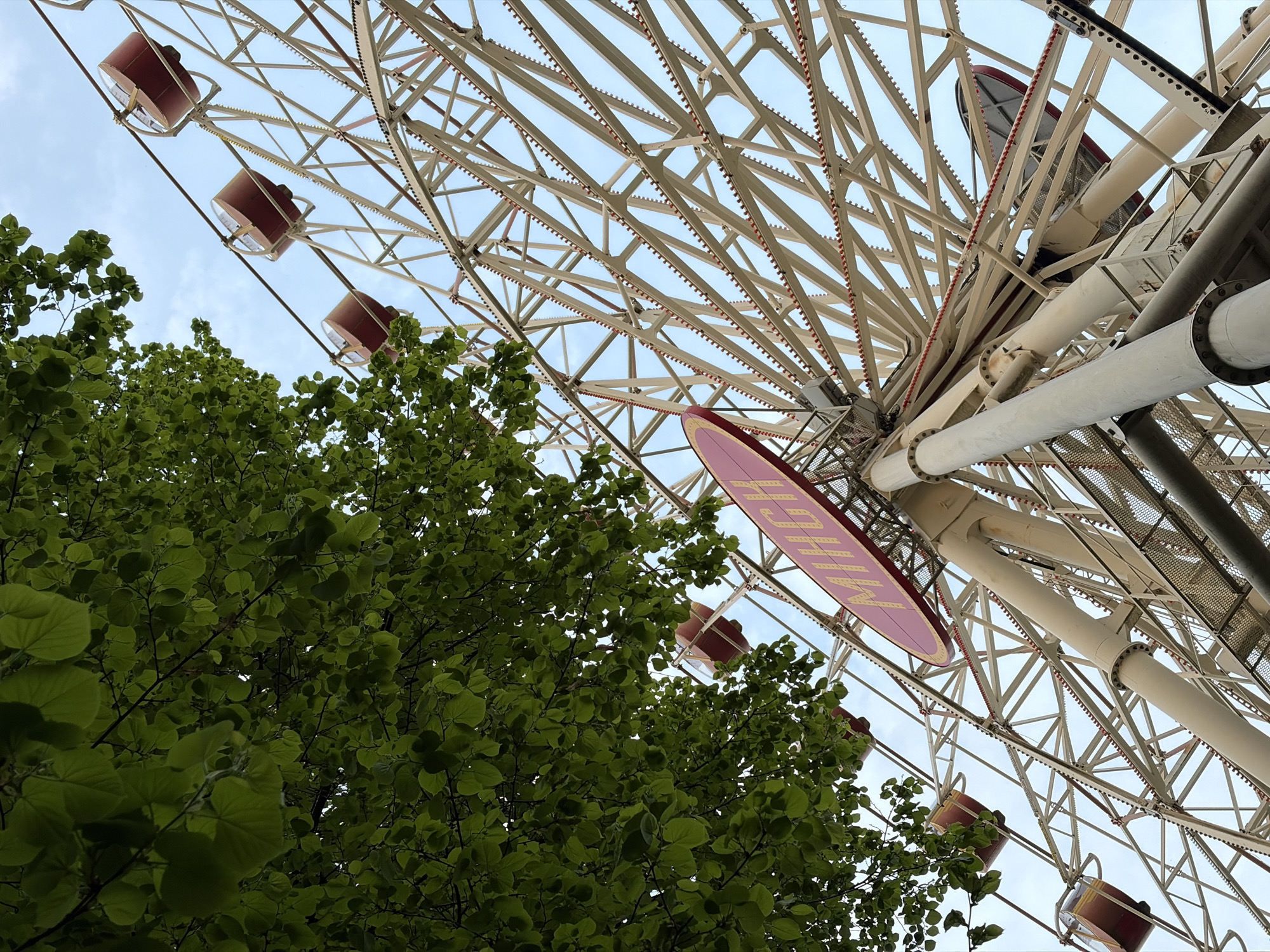 A low-angle shot captures a Ferris wheel and foliage against a light sky, located in the city of Unknown, Unknown. The Ferris wheel's white structural beams, decorated with small red lights, radiate outwards. Passenger cars, predominantly brown, are attached to the wheel's outer circumference. Dense green foliage fills the lower left corner. A red sign attached to the Ferris wheel is visible.