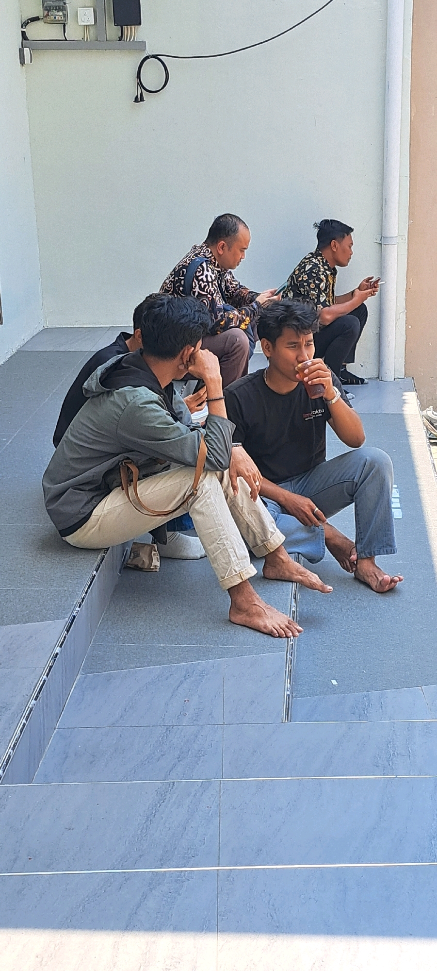 Four men are seated on outdoor steps. Two men in the foreground are sitting with their legs extended, one drinking from a cup. The other two men are seated further back, one looking at his phone. The men are dressed casually, with the exception of two men in the background wearing patterned shirts. The environment appears to be an outdoor area with a light-colored wall and tiled flooring. There is electrical equipment mounted on the wall. The image was taken in Sumber Gedang, Indonesia.