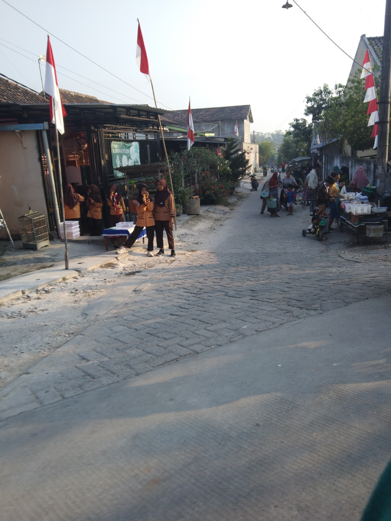 The image depicts a street scene in Klepek, Indonesia, likely during the daytime. The street is paved with cobblestones and slopes gently upwards into the distance. On either side of the street are buildings with tiled roofs. Indonesian flags, red and white, are prominently displayed, attached to poles and strung across some areas.
In the foreground on the left, several individuals are gathered near a building entrance. Two young women, possibly students in uniform, are standing on the paved walkway. One is holding a small object, perhaps food, and looking towards the camera. Behind them, more individuals are visible through an open doorway. A birdcage is also present.
Further down the street, a market appears to be set up. Several people are browsing or interacting with vendors. There are stalls with goods displayed, and some individuals are pushing carts or riding bicycles. The atmosphere suggests a local gathering or market day. The overall environment is urban with a mix of residential and commercial elements. The lighting indicates it is either early morning or late afternoon.