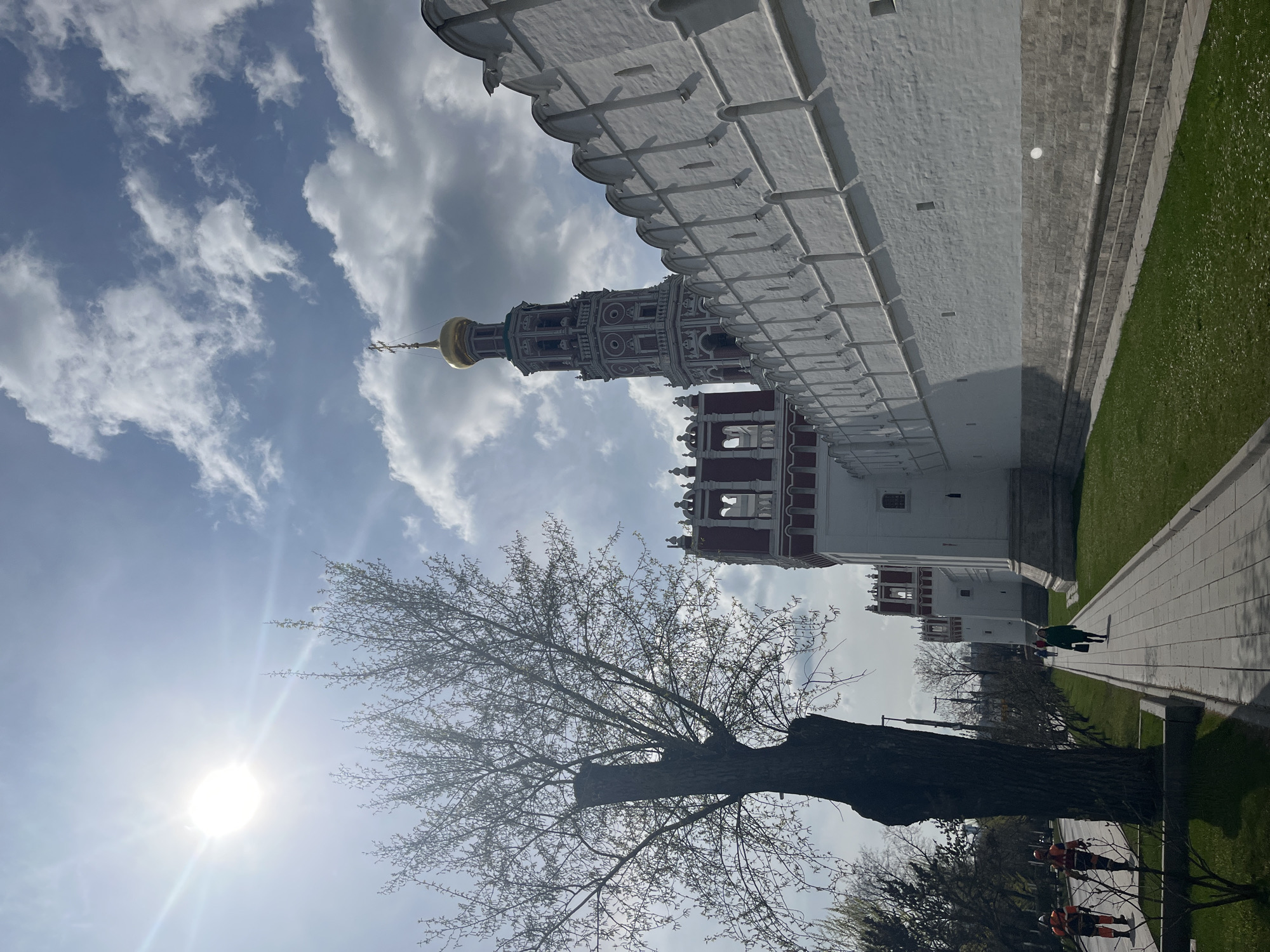 The image captures a view of the Novodevichy Convent in Moscow, Russia. A tall, ornate bell tower with a golden dome rises against a partly cloudy sky, partially obscured by the branches of a leafless tree in the foreground. A long white wall with crenellations runs along the right side of the frame, partially shadowing a paved walkway and a grassy area. Two figures are visible on the walkway in the middle ground, with additional people faintly discernible further in the distance. In the bottom foreground, the trunk of a large tree is visible, along with some shrubs. Below the tree, a team of tree trimmers, are visible working along the side. The sun shines brightly in the sky, casting a glare.