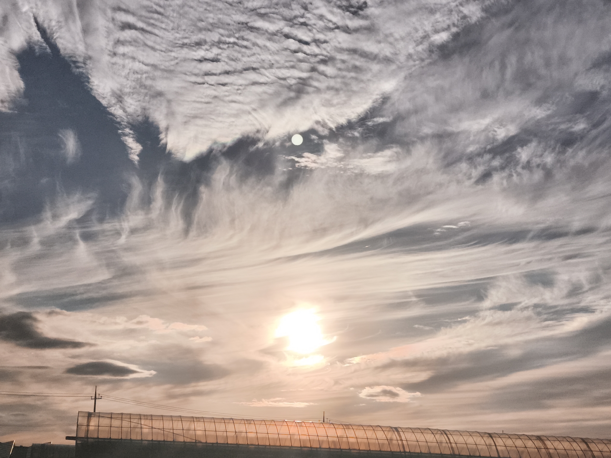 The image shows a sky filled with dramatic, wispy clouds, typical of a sunset or sunrise. The sun is visible in the lower half of the image, appearing as a bright, glowing orb, tinged with orange and pink hues from the atmospheric scattering of light. Higher in the sky, a faint circle, possibly the moon, is partially obscured by clouds. In the foreground, a long, semi-cylindrical structure, likely a greenhouse or tunnel, spans horizontally across the bottom of the frame. This structure has a translucent, ribbed surface and appears to be illuminated from within or by the fading sunlight. A solitary utility pole with wires is visible above the structure, suggesting an outdoor, possibly rural or industrial, setting within Asan-si, South Korea. The overall impression is one of a serene, yet dynamic, natural phenomenon juxtaposed with human-made structures.