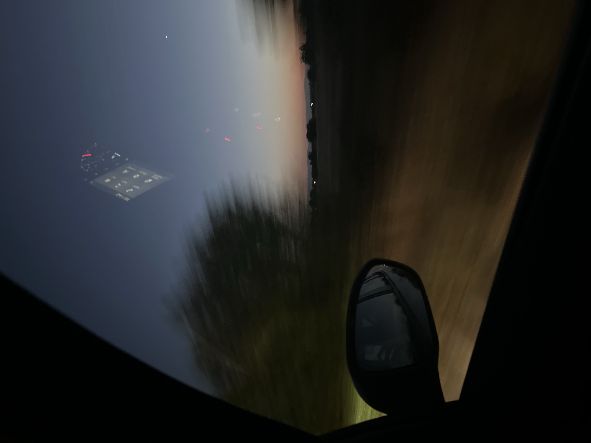 The image depicts a view from inside a moving vehicle at dusk or dawn. The foreground is dominated by the dark interior of the car, including the edge of the window frame and a side-view mirror. The mirror reflects a blurry image of the car's interior, including what appears to be a dashboard with illuminated gauges, suggesting the car is in motion.
The outdoor scenery is blurred due to the vehicle's movement, creating a sense of speed. On the left side of the image, blurred green foliage is visible, likely trees or bushes alongside the road. The sky transitions from a deep blue at the top to softer hues of orange and pink near the horizon, indicating either sunrise or sunset.
In the distance, a dimly lit landscape with silhouetted trees and possibly some faint lights from structures can be observed. There are also a few blurry red and white lights visible in the sky, which could be aircraft lights or other atmospheric phenomena.
The location is described as Skutare, Bulgaria. This implies the scene is set in or around this Bulgarian locality. The overall mood is serene and transient, capturing a moment of travel during twilight.