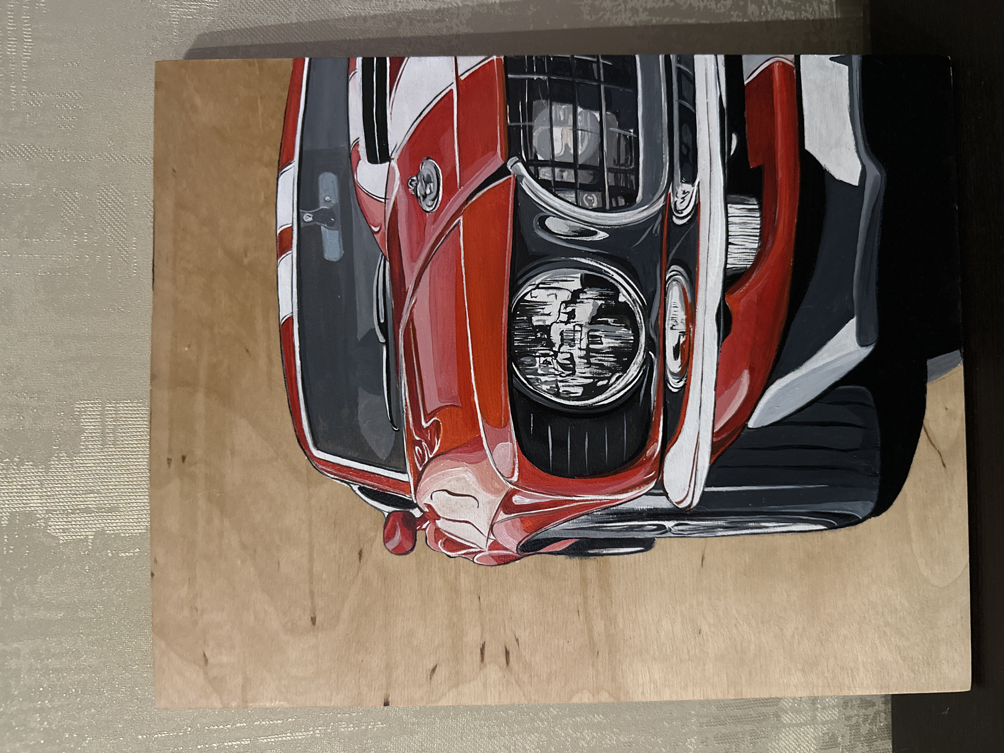 An artistic rendering of a vintage red muscle car, likely a Ford Mustang, is depicted on a light-colored wooden panel. The car is shown from a partial front three-quarter view, with its left side and front fender prominently featured. It has white racing stripes extending over the hood and roofline. The headlights are round and detailed, and the grille is a classic mesh pattern. The painting utilizes a realistic style with strong highlights and shadows, emphasizing the car's curves and metallic sheen. The background of the wooden panel is a warm, natural wood grain, with a textured, light-colored material visible behind the panel, suggesting it might be leaning against a wall. The image is a close-up of the artwork itself, with no indication of a specific city or location beyond the artwork.